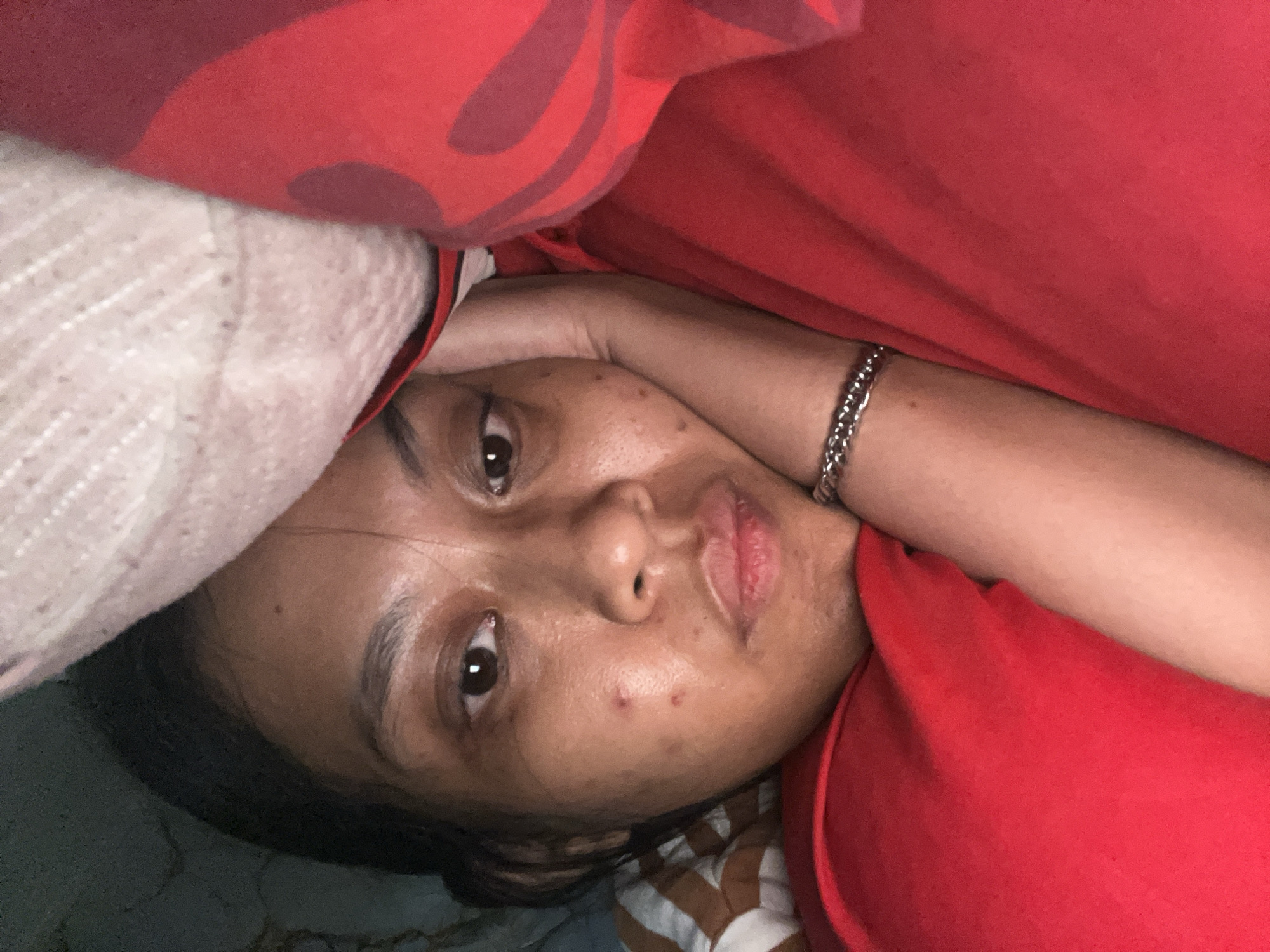 The image is a close-up, low-angle selfie of a young South Asian woman lying down, with her head propped on her arm and a pillow. She is wearing a red shirt and a silver bracelet. Her face is turned towards the camera, and she is looking directly at it with a gentle expression. The background is out of focus, but appears to be a bedroom with patterned bedding. The overall lighting is dim, suggesting it is either nighttime or indoors with low light. There are no discernible elements in the image that would indicate a specific location in Kecamatan Pati, Indonesia.
