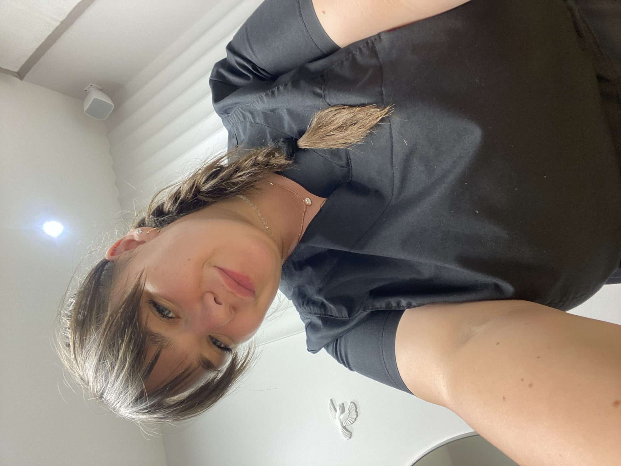 A first-person perspective shot captures a woman from a low angle. She is wearing a black scrub top and has her hair in a braid that hangs over her shoulder. A delicate necklace with a small heart pendant is visible around her neck. Her face is angled towards the camera, and she appears to be smiling faintly, with her eyes looking slightly to the side. The background is a plain white wall with a subtle textured pattern, possibly indicating a ceiling or a wall with paneling. A white speaker is mounted in the upper left corner of the frame, and a small, white decorative bird figurine is visible on the wall to the right, closer to the bottom of the image. The lighting suggests an indoor setting, possibly a room with artificial light sources, as a bright circular light is visible on the wall to the left. The overall impression is informal and personal, perhaps a selfie taken in a home or a professional environment. There is no visible text in the image.
