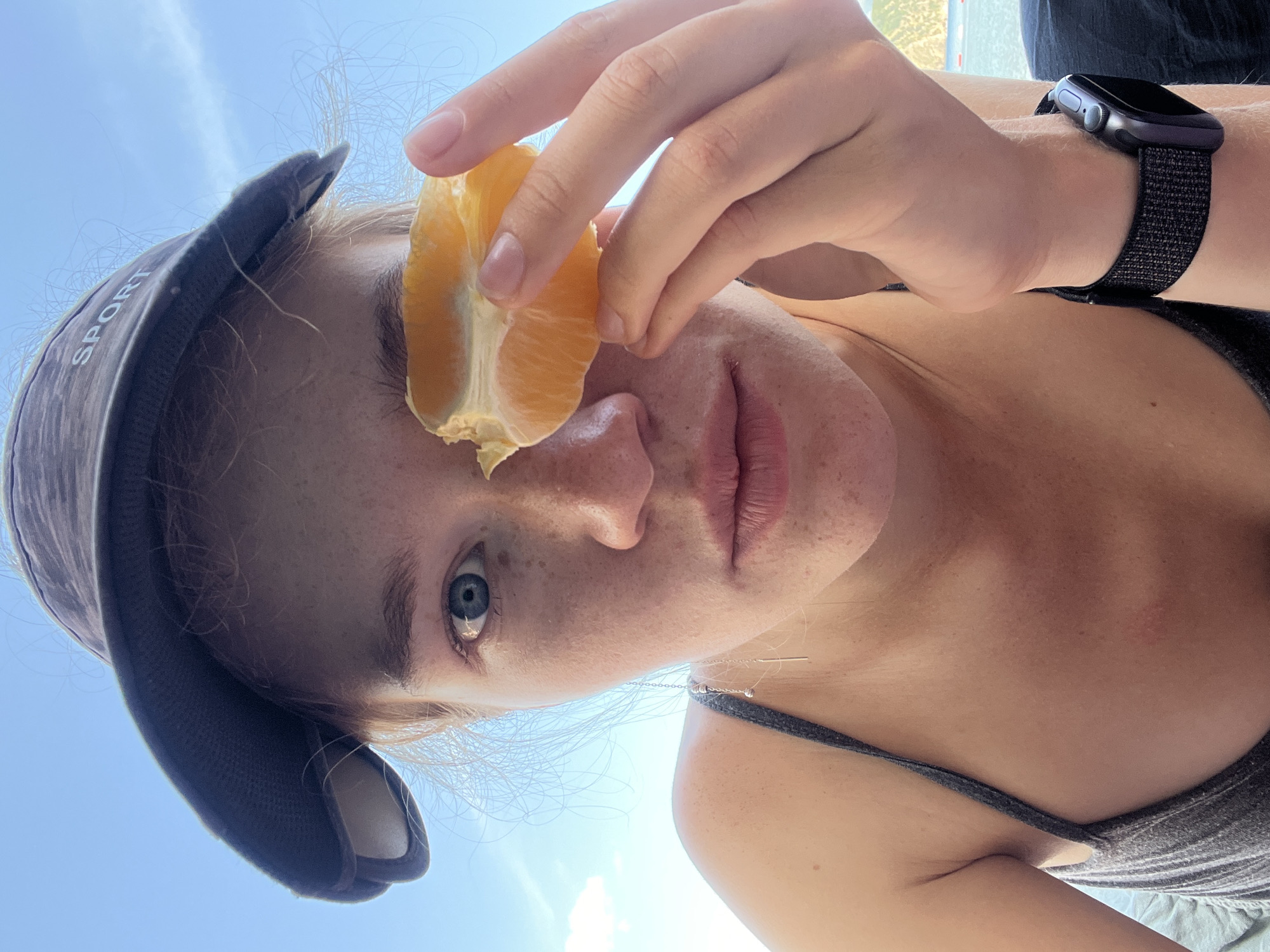 A young woman holds an orange slice to her eye, obscuring it. She wears a baseball cap and a watch on her wrist. The photograph is taken from a low angle, with the sky visible in the background. The location is possibly in Budva, Montenegro, although there are no specific details to confirm it.