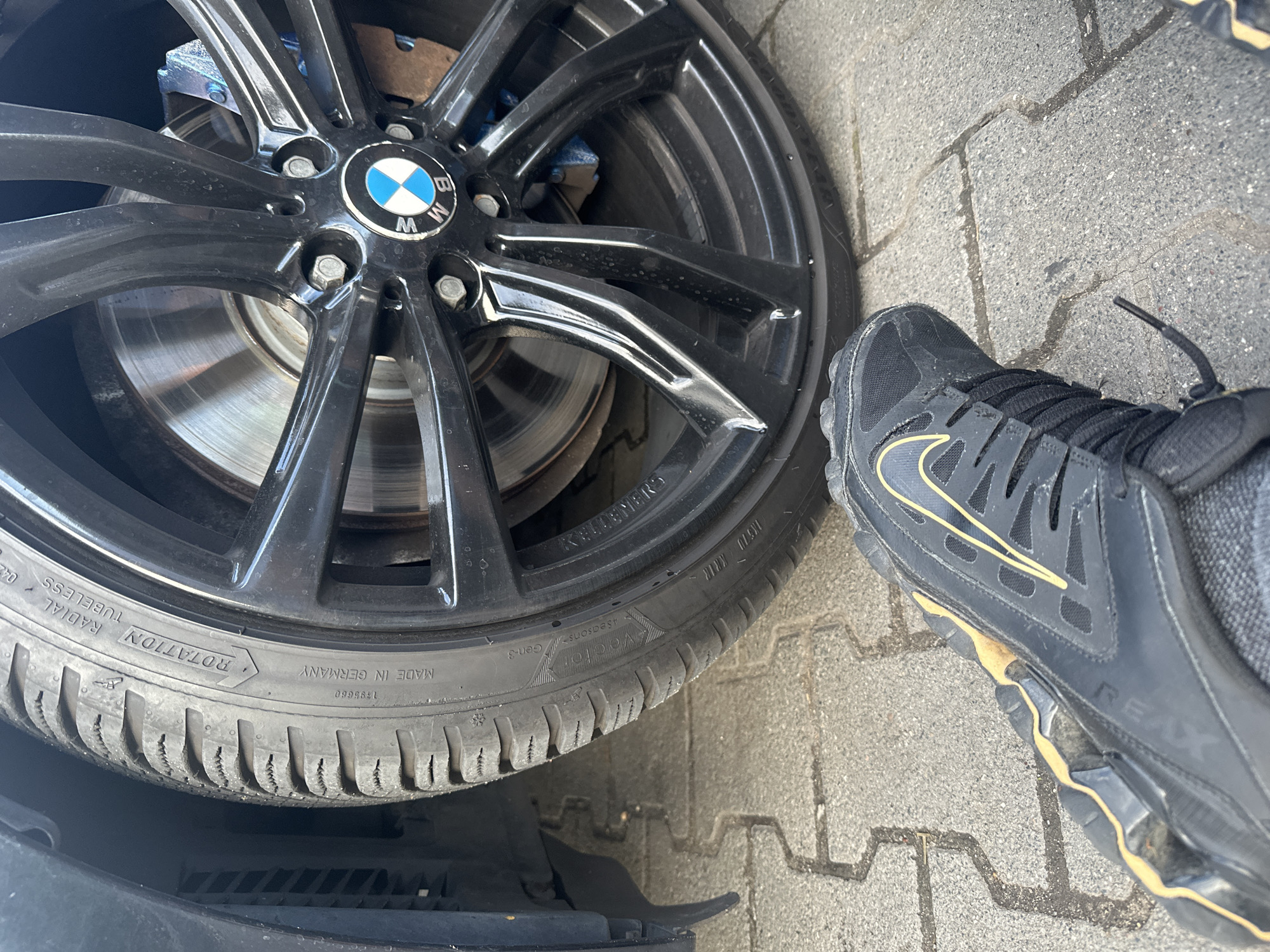 A low-angle, close-up shot shows a person's left foot wearing a black and gold Nike sneaker and the front wheel of a car with a black BMW rim and a tire with "MADE IN GERMANY" visible. The car wheel is on a paved ground with interlocked rectangular stones. The shoe is angled towards the car wheel.