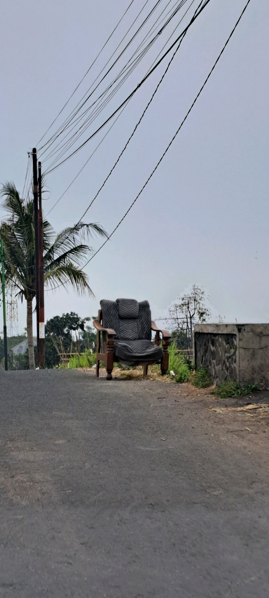 An old, dark upholstered armchair with wooden armrests and legs sits on an asphalt road in Ketanireng, Indonesia. The armchair is positioned facing the camera, slightly off-center to the right. In the background, a coconut palm tree stands to the left of the armchair, beside a utility pole supporting numerous power lines that crisscross the sky. Beyond the palm tree and armchair, there is lush green vegetation, including tall grasses and trees, under a hazy, overcast sky. To the right of the armchair, a low concrete wall with a rough stone facade runs parallel to the road, bordered by more grass and vegetation. The scene is outdoors, suggesting a rural or semi-rural setting. There are no people or animals visible in the image.