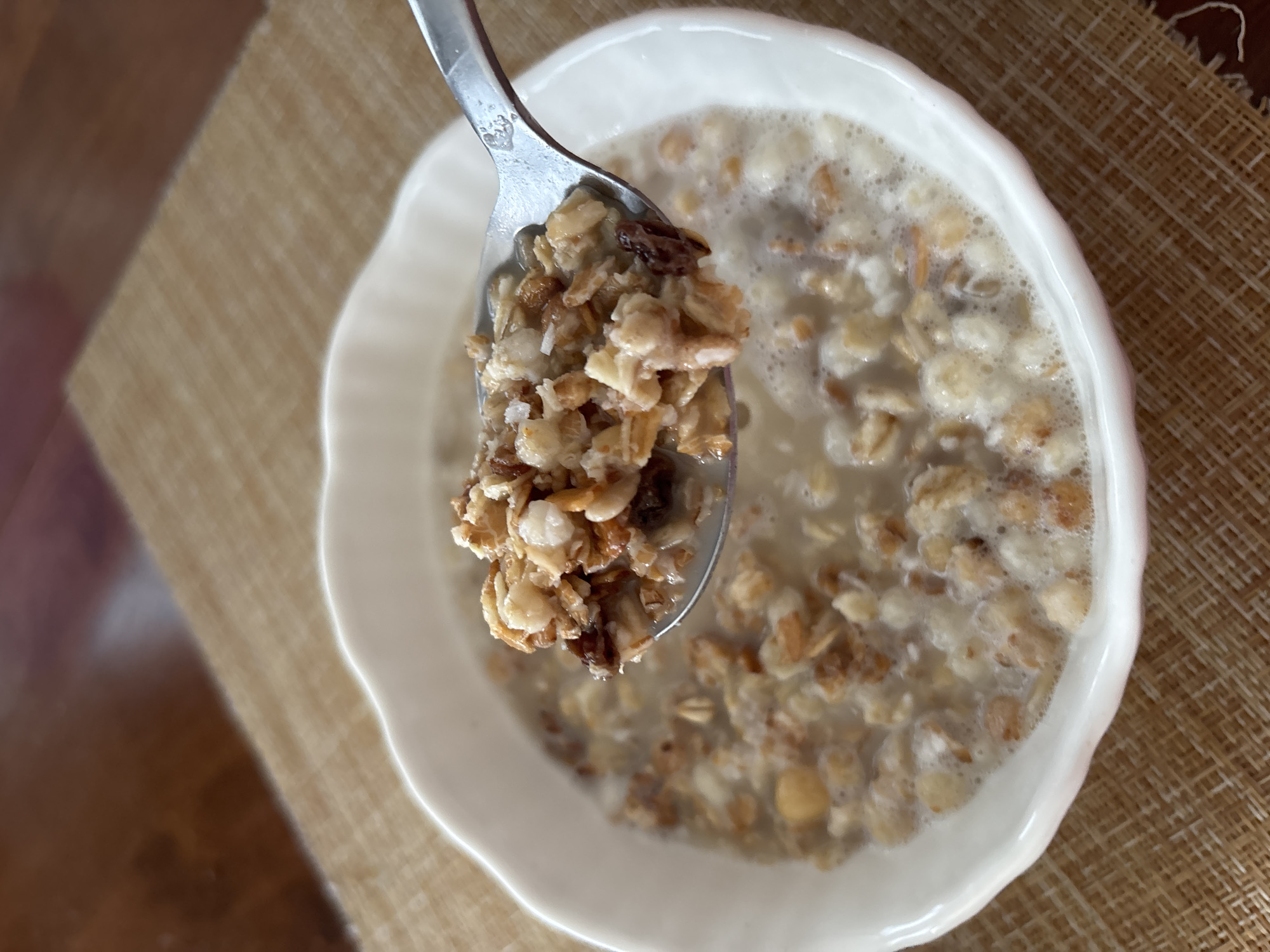 A close-up, eye-level shot shows a white, scalloped-edge bowl filled with granola and milk on a woven placemat. A spoon, held by an unseen person, is scooping up a portion of the granola from the bowl, which appears to be in the process of being eaten. The background is blurred, showing a wooden surface. There are no visible indications of the city of Sochi, Russia, or any specific environmental details to determine its location.