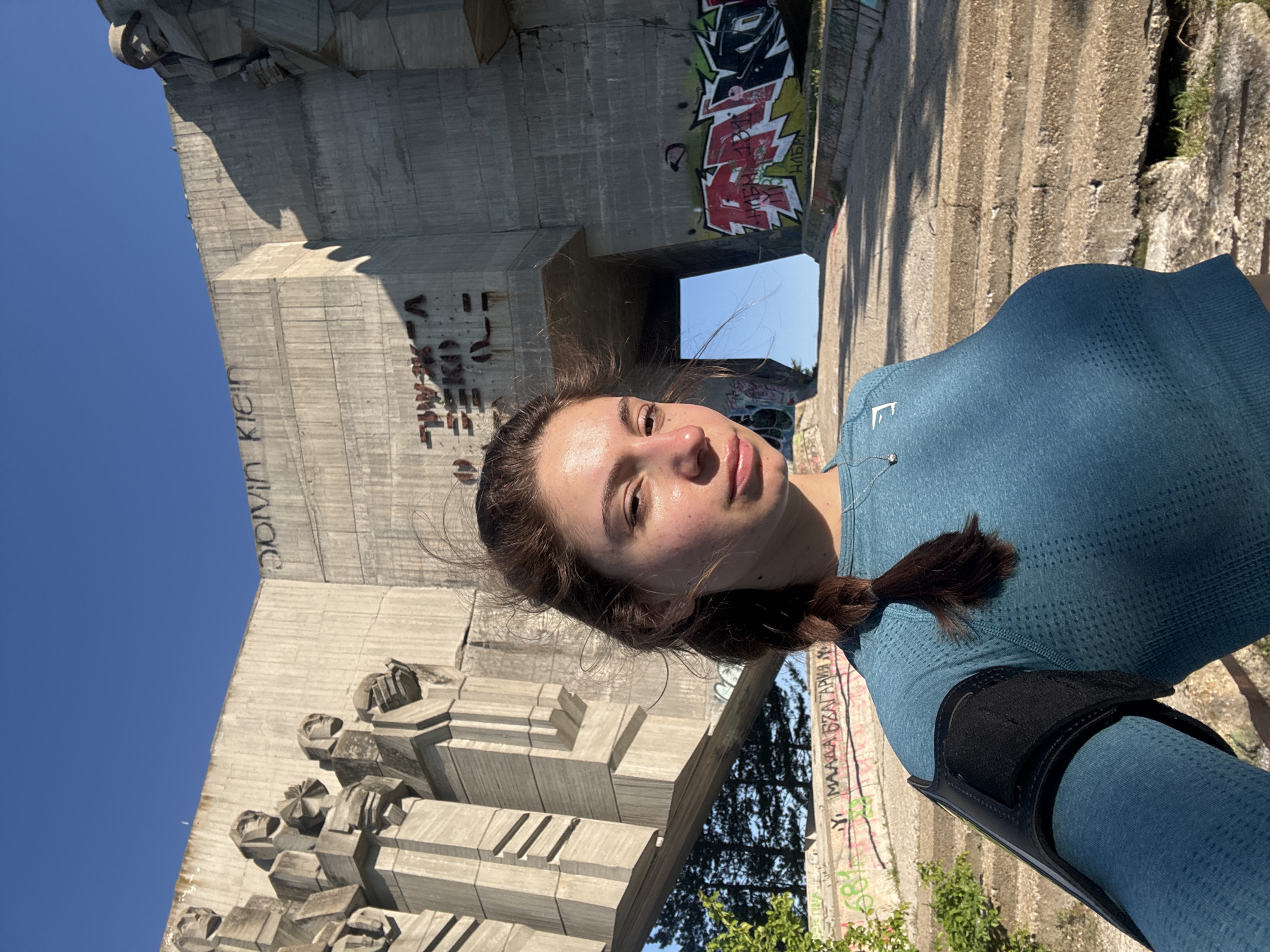 A woman poses for a selfie in front of the Buzludzha Monument in Varna, Bulgaria. The monument is large and concrete, with some graffiti on the walls. The sky is blue. The woman is wearing a blue athletic top and has a black arm band on her arm.