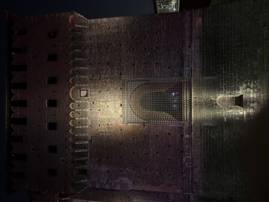 The image shows a section of a brick building, possibly a castle, at night. Two spotlights illuminate the weathered stone facade, emphasizing its texture and architecture. A decorative crenellation is visible on the left side of the building. A large, arched gate is caged in metal netting.