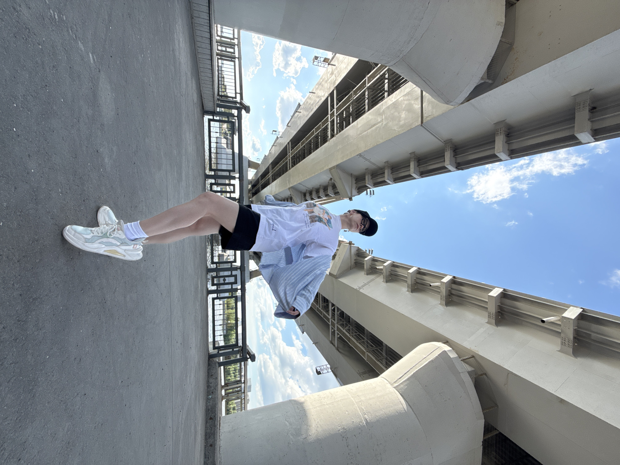 A person is standing on a concrete surface next to a metal railing in Kazan, Russia, with a view of a bridge structure and a partially cloudy blue sky. The person is wearing a black cap, white t-shirt, black shorts, a light blue jacket, white socks, and white sneakers. The bridge structure is made of concrete and metal, with a network of beams and supports visible. The person is positioned diagonally in the frame. The ground is paved and shows signs of wear.