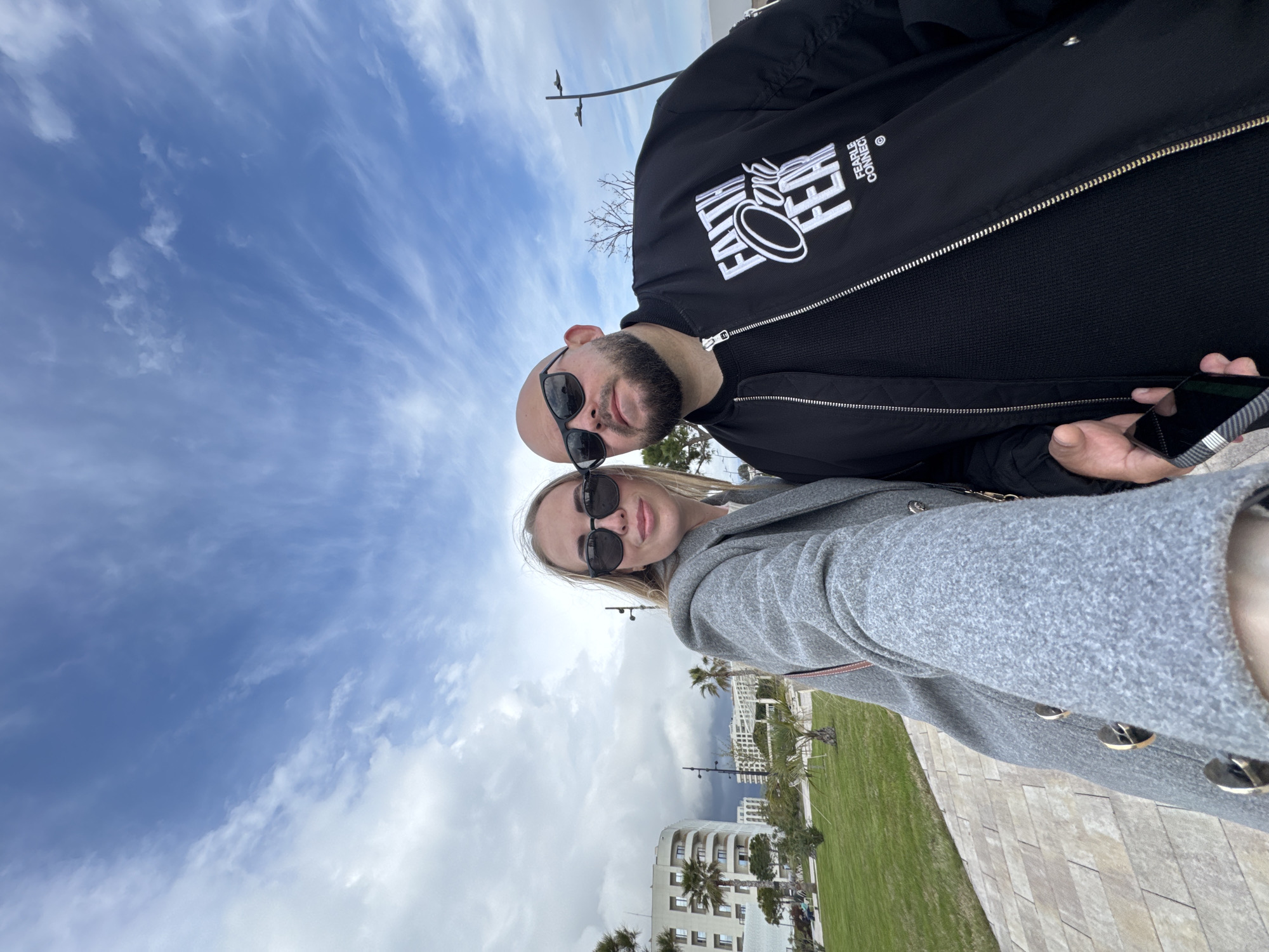 A selfie taken outside in what appears to be the city of Sirius, Russia. A man with a bald head, beard, and sunglasses is on the top right, wearing a black jacket. A woman with blonde hair and sunglasses is on the bottom left, wearing a gray coat. In the background are buildings, grass, and palm trees, suggesting a warm climate. The sky is partly cloudy.