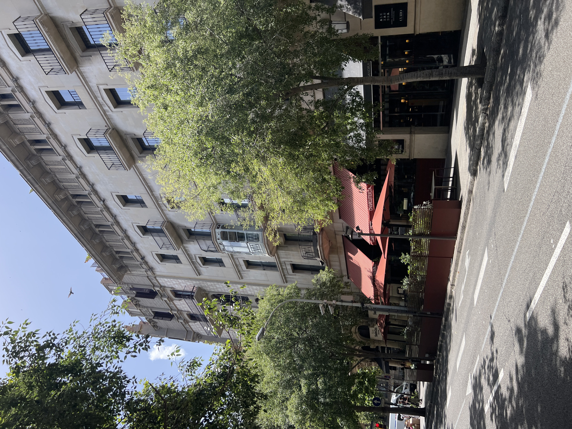 The image shows a street scene in Barcelona, Spain, on a sunny day. The focal point is a multi-story building with a cream-colored facade, featuring several balconies with black iron railings. The lower floors of the building house shops or restaurants, indicated by awnings and window displays. Two large red awnings are prominent, one with the word "CHARCUTERIA" visible. Trees with lush green foliage line the sidewalk, casting shadows on the street. The street itself is paved and has lane markings, including white arrows. A street lamp and some security cameras are visible on a pole. A few people can be seen on the sidewalk in the distance, and a bird is flying in the sky.