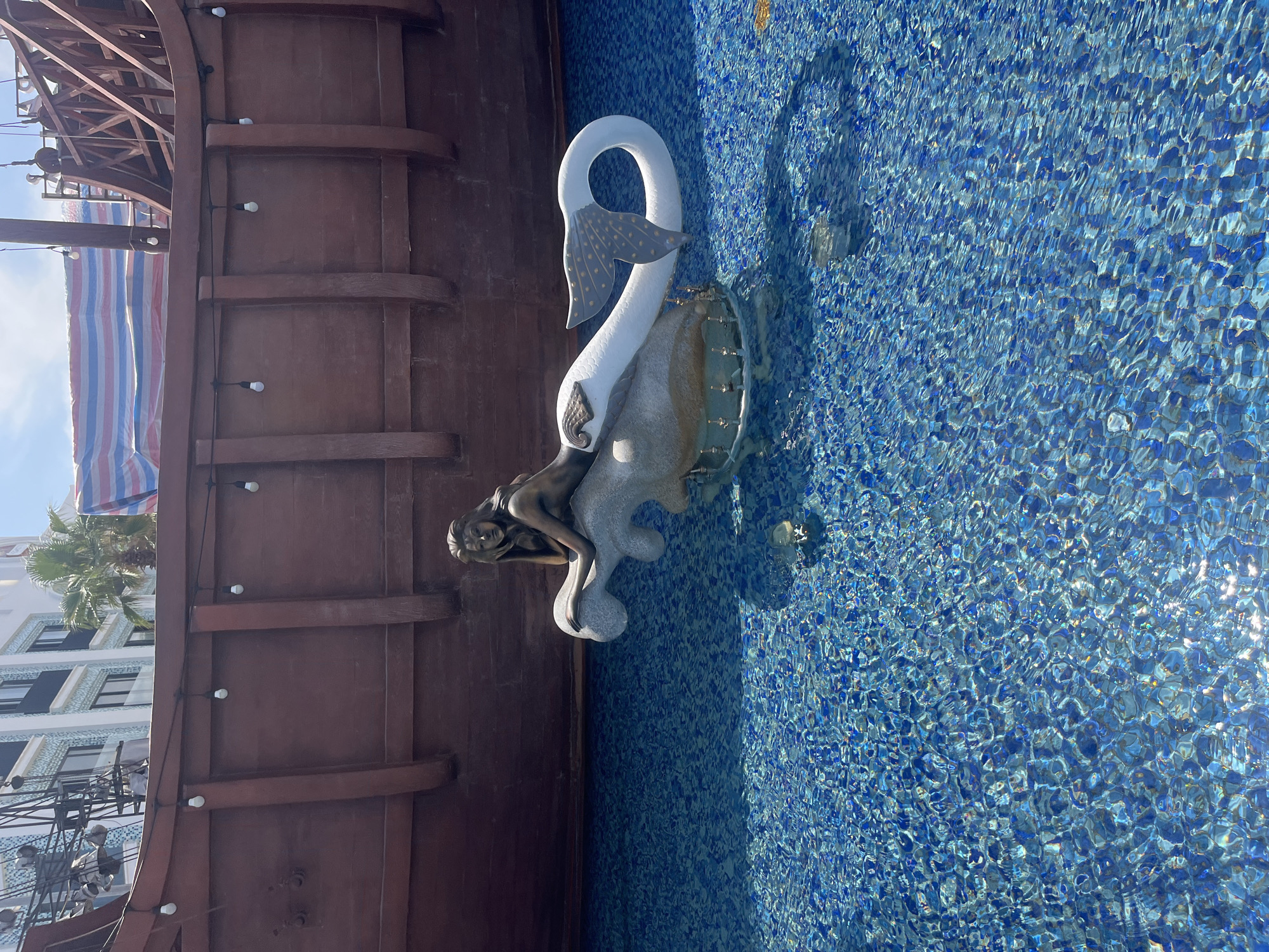 A vertical, eye-level shot shows an outdoor mermaid sculpture in a blue-tiled pool in the Bac Natural Heritage Area Trang, Vietnam. The statue features a bronze-colored mermaid reclining on a white swan. The swan has a fan-like tail with gold polka dots. The swan sits atop a light-gray stone base. The pool is made of varying shades of blue tiles. To the left, is the brown wooden structure of a ship. Strings of lights are attached to the ship structure. The backdrop shows palm trees and a white building with dark-tinted windows.