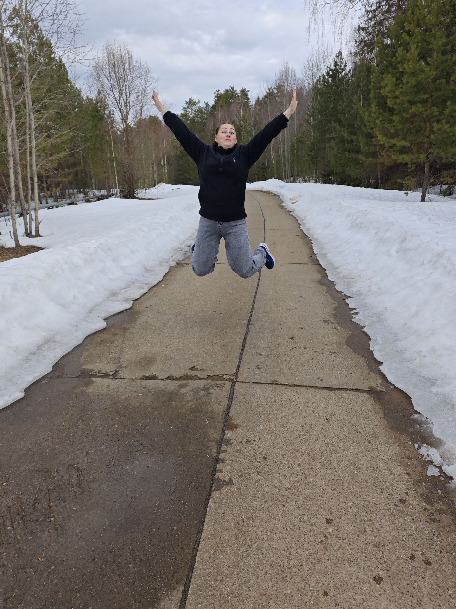 The image shows a person jumping in the air on a concrete path in a park-like setting. The person is wearing a black sweatshirt and gray jeans. They have both arms raised above their head. On either side of the path is melting snow, with trees visible in the background. The sky is overcast. The location is Марковский, Russia.