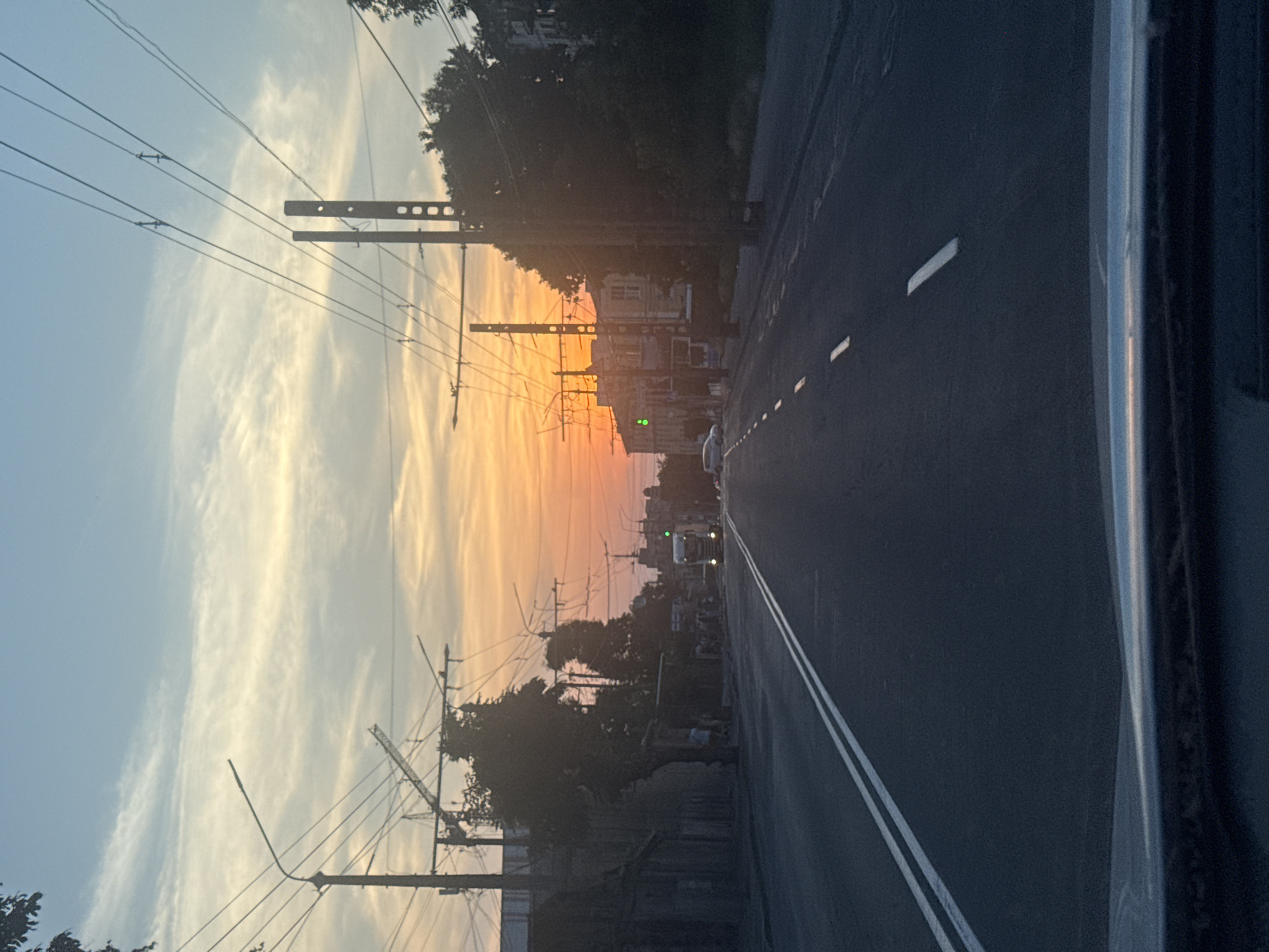 This is a vertical, eye-level shot of a street in Vinnytsia, Ukraine, during a sunset. The sky is a vibrant gradient of orange, yellow, and blue, with wispy clouds. Power lines crisscross the frame, supported by utility poles. On the left side of the image, trees and buildings are silhouetted against the colorful sky. The right side of the image shows a dark asphalt road with double white lines. A few cars are visible on the road in the distance, with headlights on, suggesting it's evening. There are also people walking on the sidewalk. The overall mood is serene, with the warm colors of the sunset creating a peaceful atmosphere. The presence of power lines and utility poles indicates an urban or semi-urban setting.
