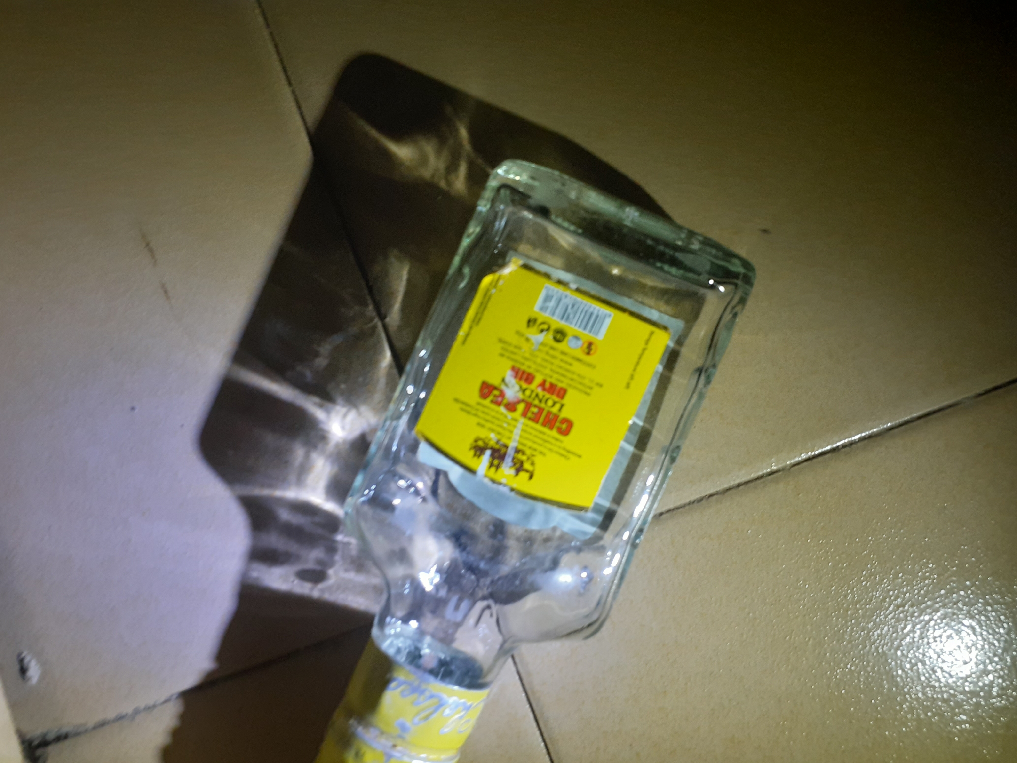 The image shows a glass bottle of Chelsea London Dry Gin lying on its side on a tiled floor. The bottle has a yellow label with black text. The background is a tiled floor with some debris. The scene appears to be indoors. There are no people or animals visible in the image. The lighting is dim, and there are shadows cast by the bottle and other unseen objects. The specific location within Jalingo, Nigeria, cannot be determined from the image alone.