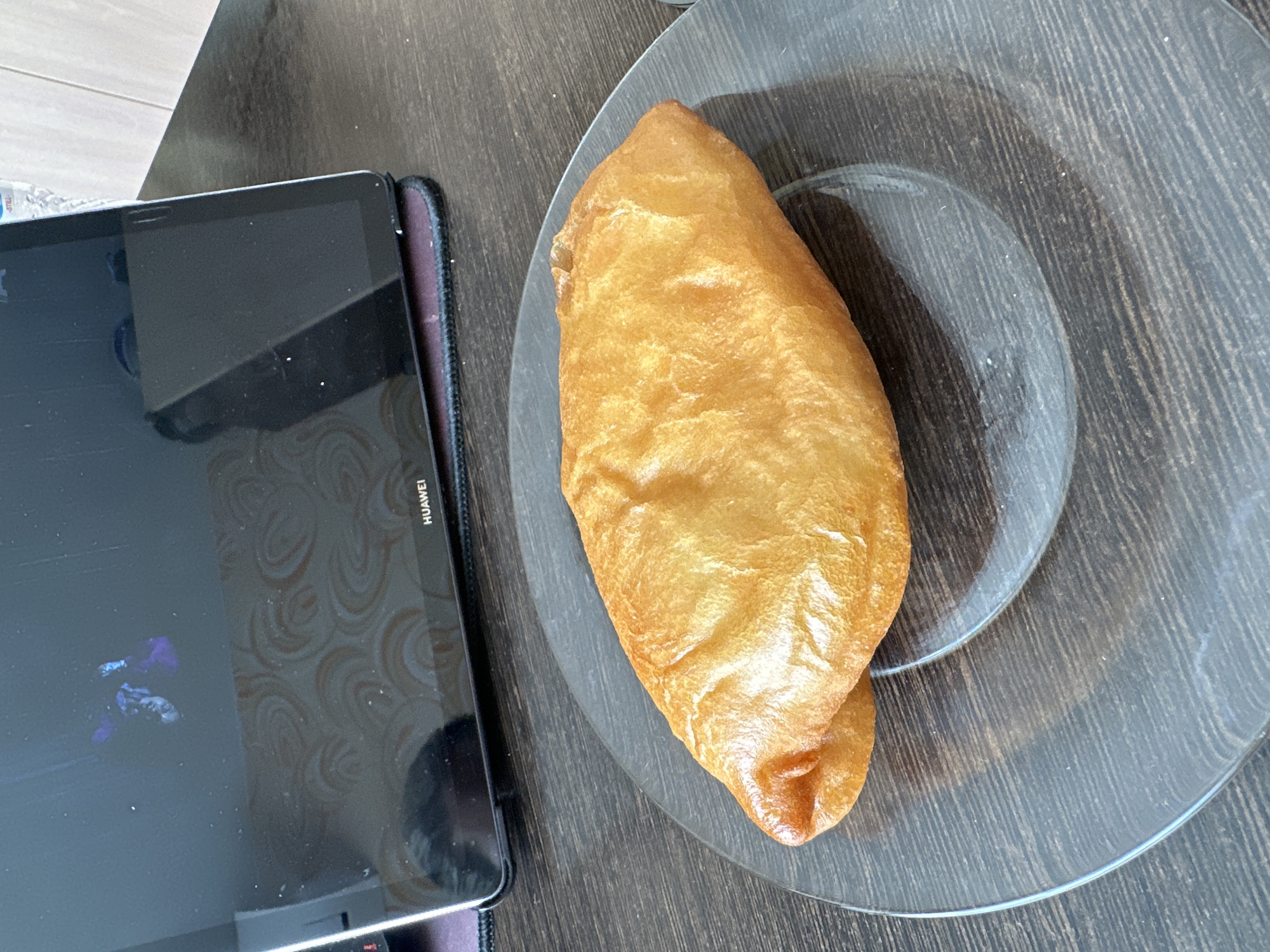 A close-up, overhead shot shows a golden-brown, fried pastry on a dark, translucent plate. The pastry is plump and appears to be filled, with a slightly uneven, puffy surface. The plate has a subtle wood-grain texture beneath it, which is also visible on the dark wooden tabletop. In the background, a dark tablet device is propped up, displaying a swirling pattern. The overall scene is indoors, with the lighting suggesting it is daytime. The image does not provide specific information to determine a location within Novoaltaysk, Russia.
