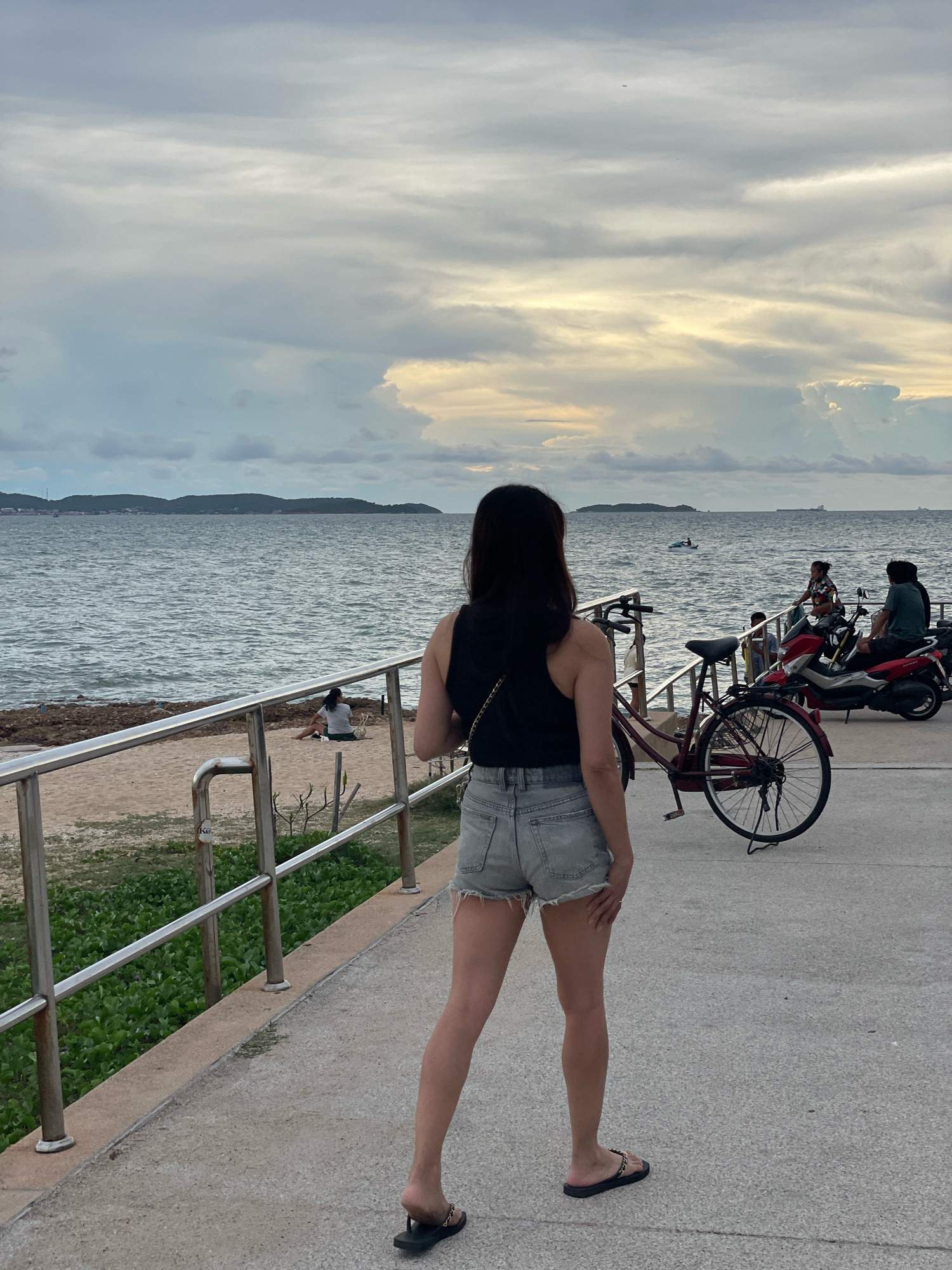 A woman stands facing away from the camera on a paved walkway along a beach in Pattaya City, Thailand. She has long, dark hair and is wearing a black tank top and light-wash denim shorts. She is wearing flip-flops. To her left is a metal railing, and beyond that, a sandy beach with some green vegetation. The sea is to the left of the beach, with a hilly coastline in the distance. On the walkway behind her, a bicycle is parked, and several people are gathered around two motorcycles. The sky is overcast with dramatic cloud formations.