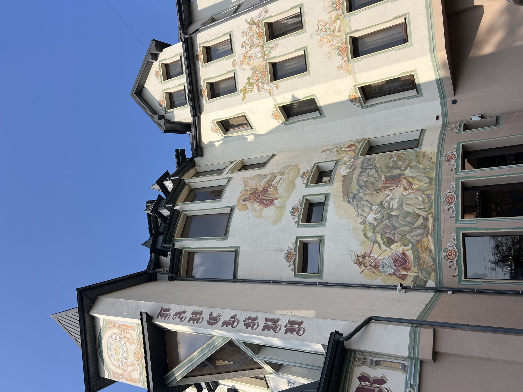 The image shows a close-up, low-angle exterior view of a building with ornate murals, likely in Yaremche, Ukraine. The building has a distinct architectural style with a clock tower on the left featuring a Roman numeral clock face and cherubic figures. Vertical lettering in a gothic script is visible on the side of the tower. The building's facade is painted in light colors and adorned with large, detailed murals depicting pastoral scenes with figures in historical attire, musicians, sheep, and mountainous landscapes. The murals are bordered by decorative floral motifs. Several windows with dark frames are visible on different levels of the building. The sky above is a clear, bright blue. The lower part of the building has a darker, possibly rose-colored base. The overall impression is of a highly decorative and possibly historical building, characteristic of some tourist destinations.