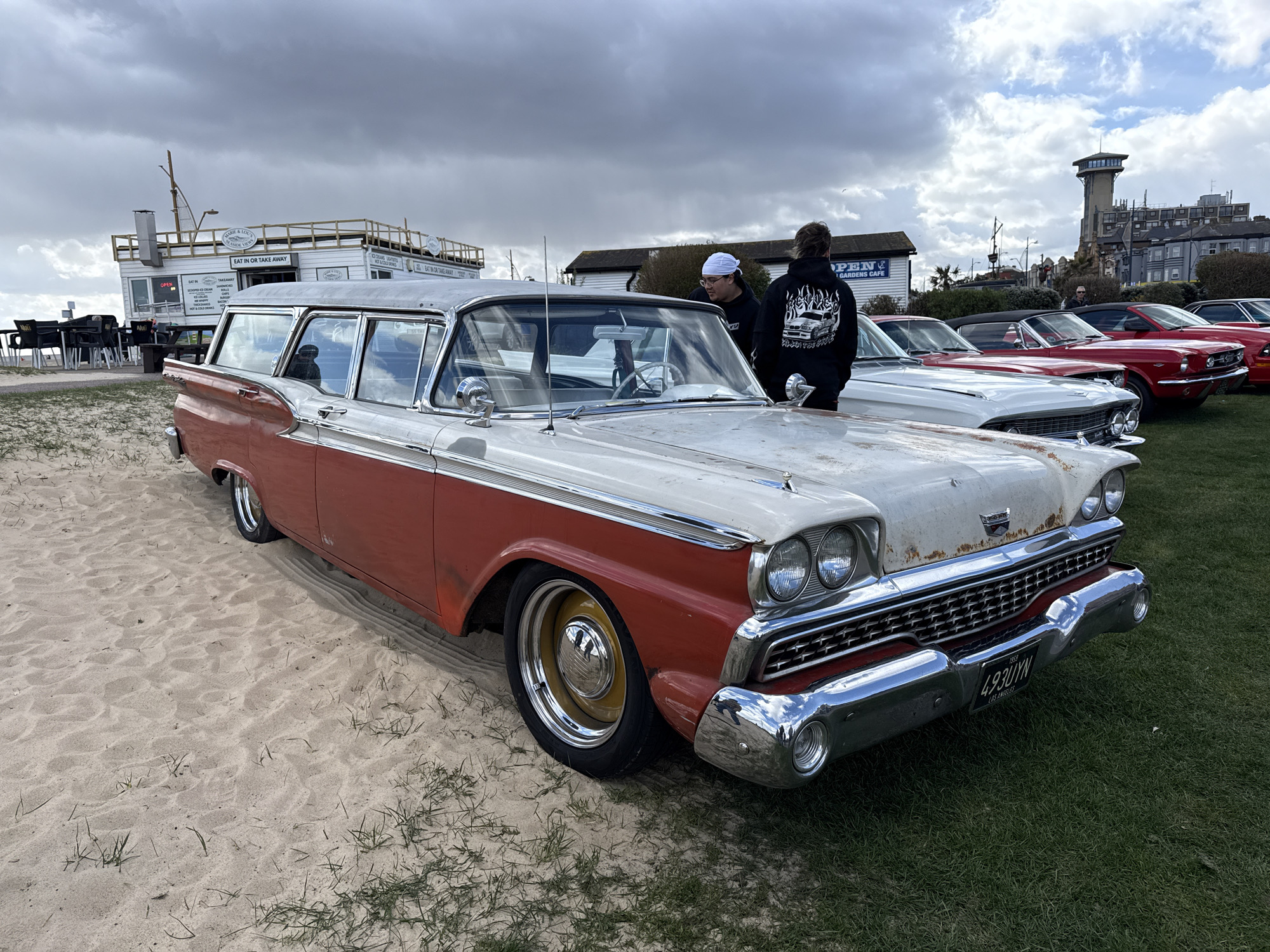 The image shows a vintage red and white station wagon parked on a patch of sand and grass, likely at a car show or gathering. The car has a license plate reading "493UYN" and a "1959 LOS ANGELES" plate frame. Two people are standing near the car, one wearing a white cap and another wearing a black hoodie with a car graphic on the back. Several other classic cars are parked in a line behind the station wagon, including red and white convertibles. In the background, there is a building labeled "EAT IN OR TAKE AWAY" and "OPEN GARDENS CAFE", as well as a tall tower in the distance. The scene appears to be in Great Yarmouth, United Kingdom, judging from the architectural style of the buildings and the cars' license plate. The sky is overcast and cloudy.