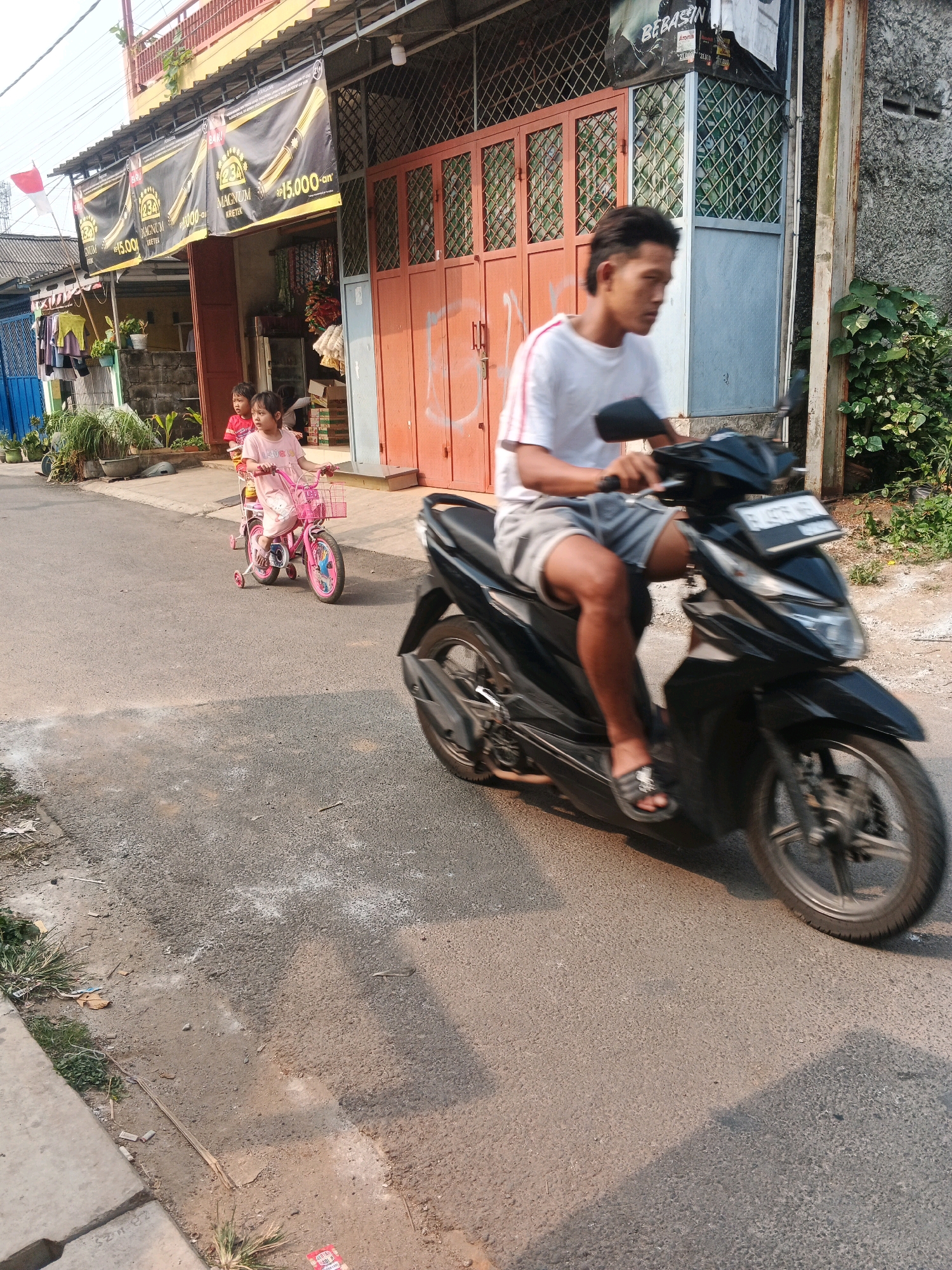 In Kadu, Indonesia, a man rides a black motorcycle down a paved road. He is wearing a white t-shirt and gray shorts. The motorcycle has a license plate with black text on a white background. In the background, two young children are on a pink bicycle. The girl in front is wearing a pink shirt and the boy behind her is wearing a red shirt. The bicycle has training wheels and a basket. They are in front of a building with an orange facade and a black awning displaying advertisements. The environment appears to be a street in a residential area, with some greenery and buildings visible.