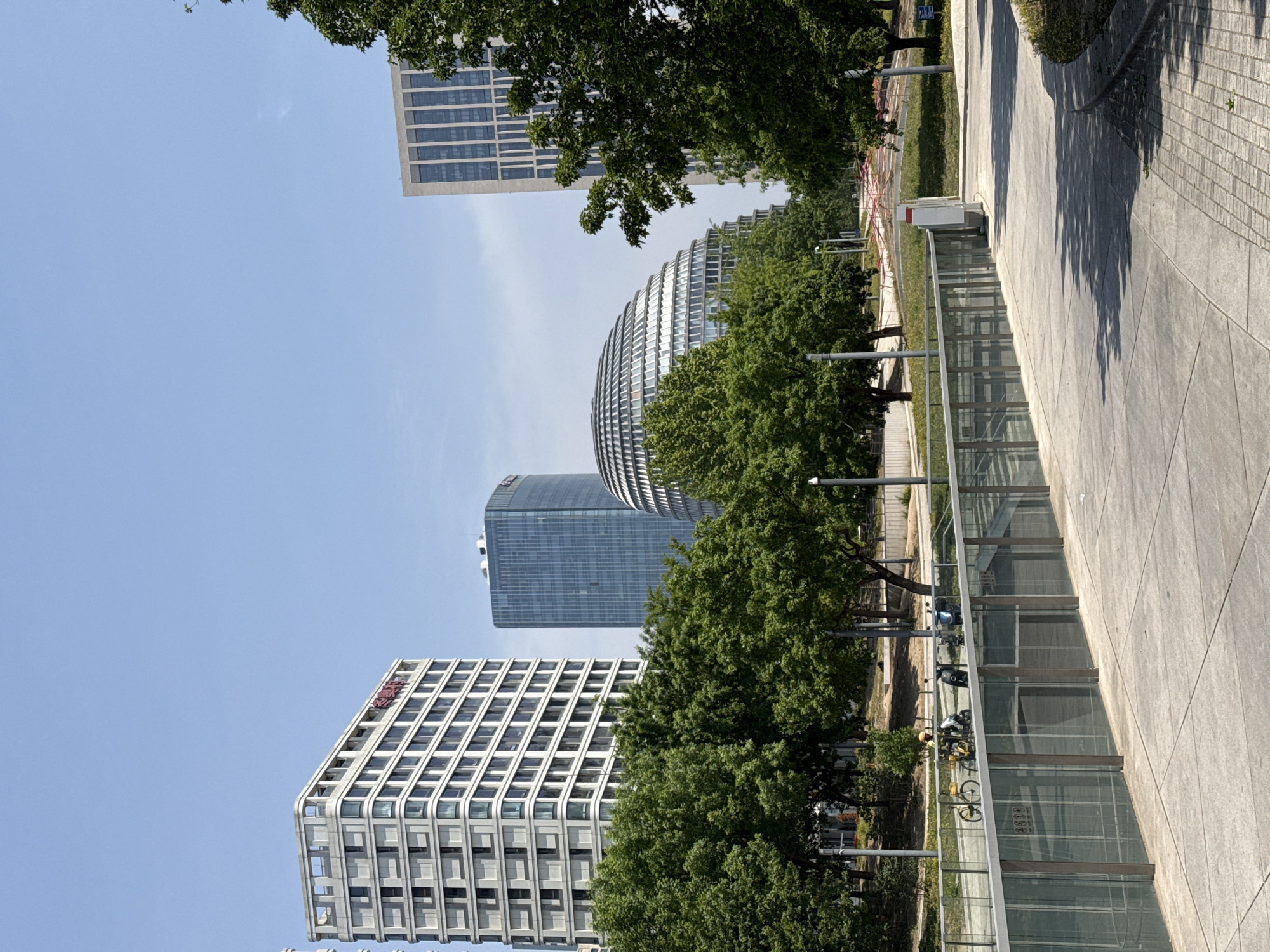 The image captures an outdoor scene in Chaoyang District, China, under a clear, blue sky. Prominent modern architectural structures, including several high-rise buildings with distinctive designs, dominate the background. The buildings feature a mix of straight lines and curvilinear forms, with glass and steel facades. Lush green trees partially obscure some of the buildings.
A pathway stretches along the right side of the frame, featuring a gray concrete surface and a glass barrier. Bicycles are parked near the glass barrier. The pathway transitions into a walkway made of brick.