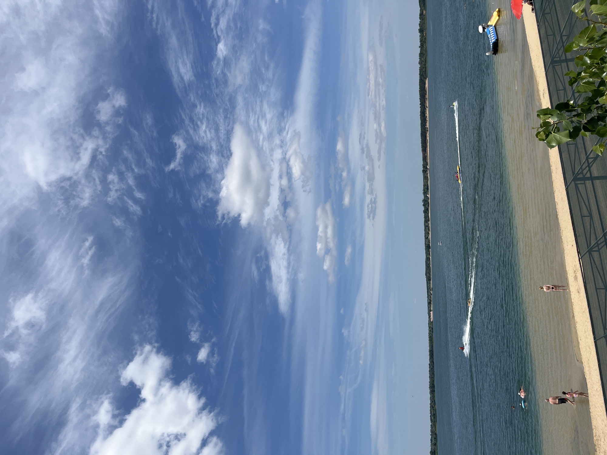 The image depicts a scenic view of a beach and lake in Yelyzavetivka, Ukraine. The sky is a vibrant blue, filled with wispy white clouds, suggesting a pleasant, sunny day. The lake, a deep teal color, stretches towards a tree-lined horizon. Several watercraft are visible on the lake, including a jet ski creating a white wake, a yellow banana boat with multiple passengers, and two inflatable water toys near the shore. A few people are enjoying the water, with some swimming and others floating on inflatables. On the sandy beach in the foreground, a few individuals are present. A man and a young girl stand in the shallow water, while a woman in a swimsuit stands on the wet sand. A fence runs along the edge of the beach, and lush green foliage from a tree drapes into the lower right corner of the frame. The overall scene conveys a sense of leisure and summer recreation.