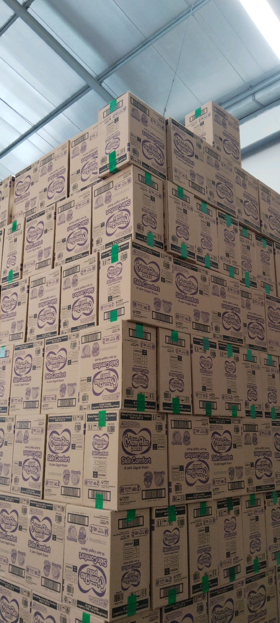 The image shows a large stack of cardboard boxes in what appears to be a warehouse or storage facility. The boxes are stacked high, filling the frame from bottom to top and left to right. They are predominantly light brown with purple and white branding and text. The branding includes the words "MamyPoko Pants" and "Skin Comfort" along with illustrations of babies and product information. Some boxes have green tape reinforcing their seams. The environment appears to be indoors, with a corrugated metal ceiling visible at the top of the image. The lighting suggests overhead artificial lighting. The image does not contain any people or animals. The scene is devoid of any obvious signs of damage or disarray, suggesting an organized storage system. The specific location within Surabaya, Indonesia, is not discernible from the visual information provided in the image itself.