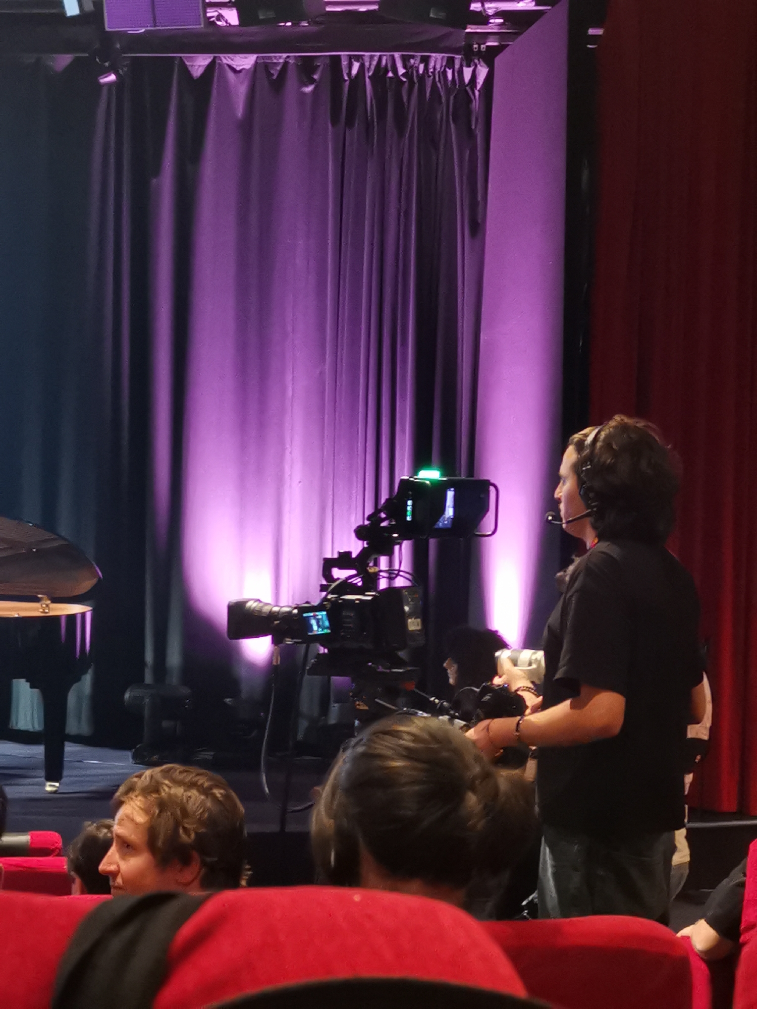 The image shows the interior of a theater, possibly during a performance or rehearsal. A black grand piano is visible on the left side of the frame, positioned on stage. A camera crew is actively working, with a professional video camera prominently positioned in the center. A crew member with a headset stands beside the camera, facing right. Several audience members are seated in the foreground. The seating is red and the stage is behind them with purple and red curtains. The location of the theater is unknown.