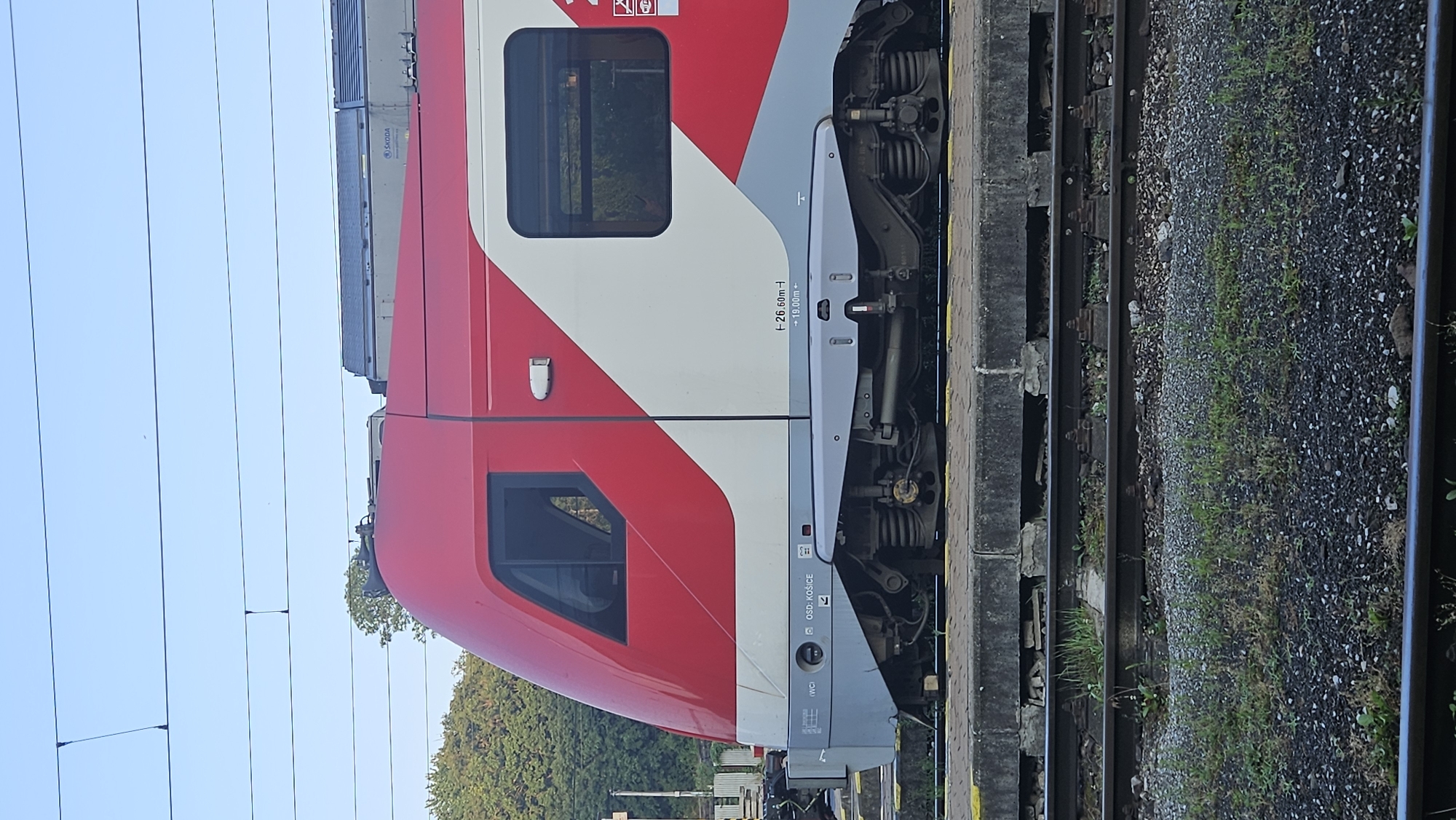 The image captures a side view of a red and white passenger train, identified by the "ÖBB KOSICE" marking on its side, at the Kysak, Slovakia train station. The train's locomotive, with its characteristic red front and white stripe, is prominently featured. The exterior of the train shows two windows, one on the front cab and another on the passenger section. Beneath the train, its wheels and suspension system are visible, resting on railway tracks. The station platform is a concrete structure, with gravel and vegetation around the tracks. In the background, trees and overhead power lines indicate an outdoor setting. The sky is clear and blue. There are no people visible in the image. The scene appears to be a static view of the train at the station, suggesting a pause in its journey.