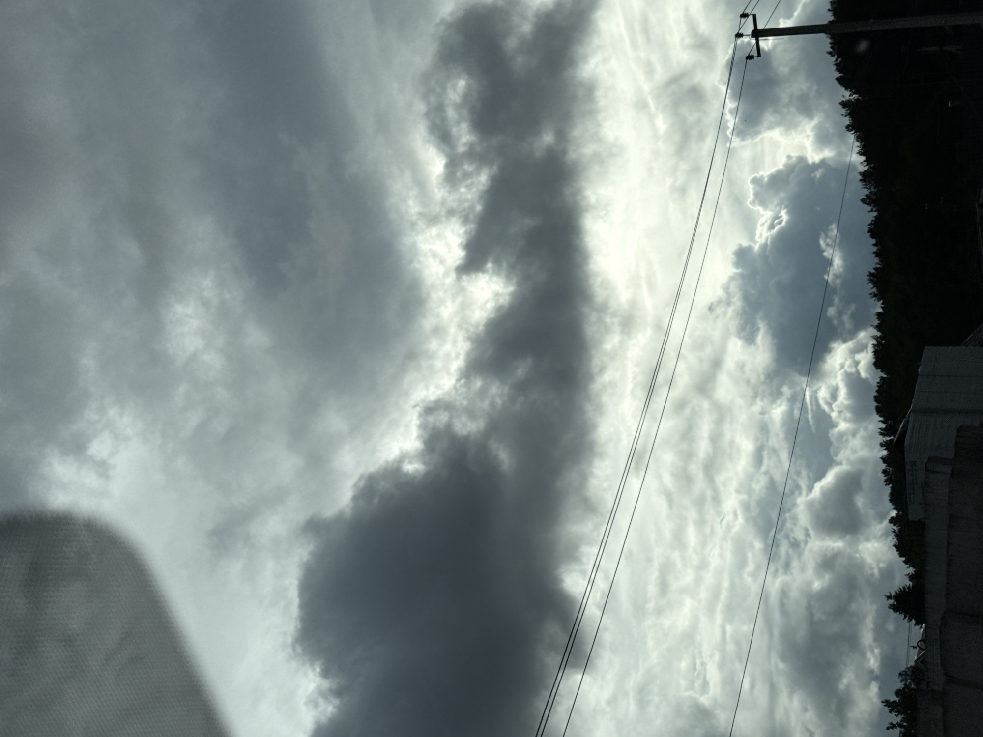 The image captures a dramatic, cloudy sky, likely viewed from inside a vehicle given the blurred reflection in the lower left corner. The dominant feature is the interplay of dark, dense clouds with brighter, luminous patches where sunlight breaks through.
A prominent, elongated dark cloud formation stretches vertically across the central part of the frame, contrasting sharply with the illuminated sky behind it. Several thin, dark utility lines or power cables traverse the sky diagonally, from the bottom left towards the upper right. These lines are supported by a dark utility pole visible on the far right side of the image.
Along the right edge, a dark silhouette of dense trees or a forest can be seen, partially obscured by the utility pole and a section of what appears to be a light-colored concrete or stone building/structure at the very bottom right. The overall impression is one of an overcast day, possibly hinting at recent or impending stormy weather, with moments of dramatic lighting as the sun peeks through gaps in the cloud cover. The time of day is not explicitly clear but the diffused light suggests either morning or late afternoon, or simply a heavily overcast period.
There is no visible text in the image. The scene is set in Chaikovsky, Russia.