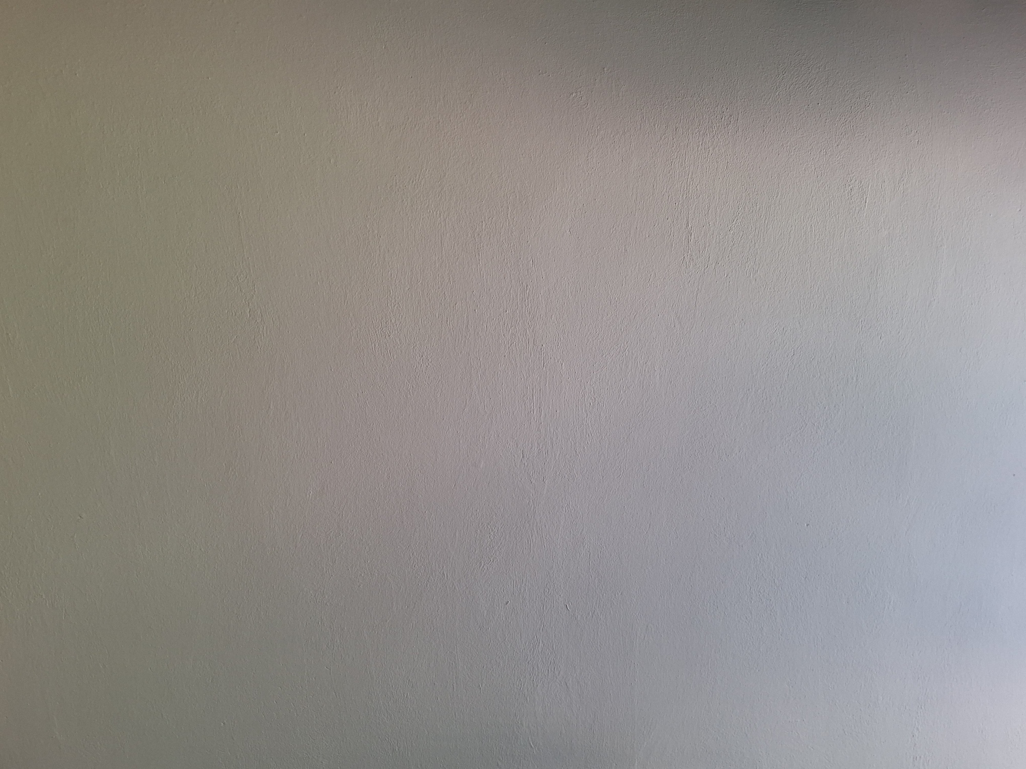 The image is a close-up, full-frame shot of a textured, off-white wall. The lighting creates a gradient effect, with the left side appearing brighter and cooler in tone, and the right side becoming warmer and slightly darker, suggesting a light source from the upper left. The texture of the wall is subtly visible, with a slightly rough, uneven surface that hints at painted plaster or stucco. There are no people, objects, or discernable features that would indicate a specific location within the city of Varna, Bulgaria. The scene is entirely abstract and focuses solely on the surface of the wall.
