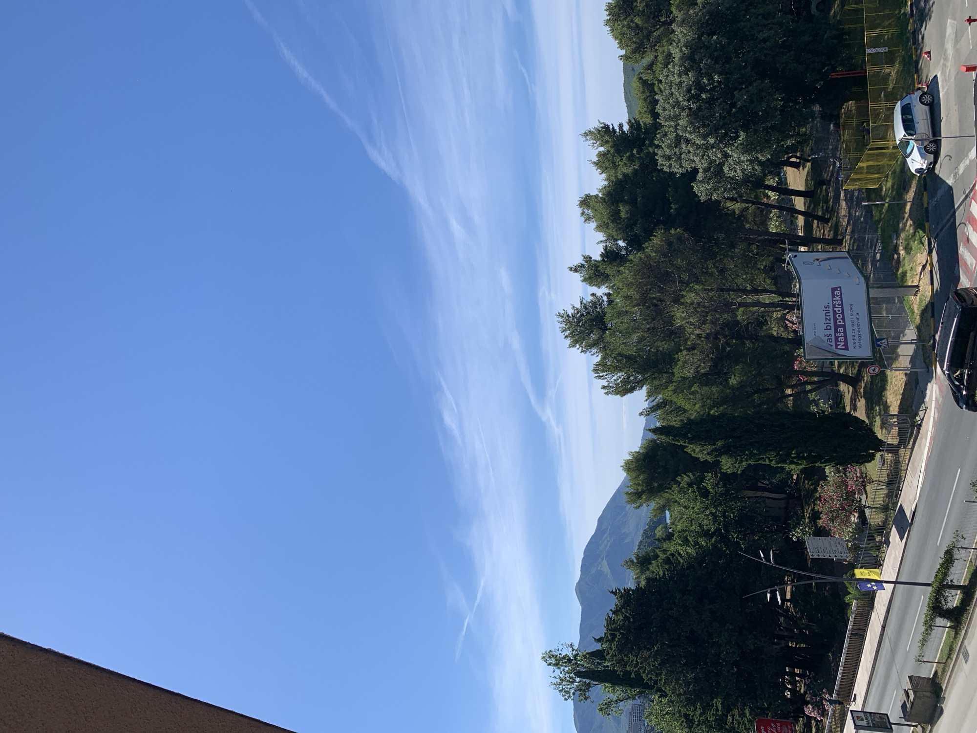 This is a ground-level view of a street in Budva, Montenegro, featuring a clear blue sky with wispy clouds and a distant mountain range. In the foreground, a paved road runs horizontally across the image, with a sidewalk and a fence bordering it. Several cars, including a silver hatchback and a black sedan, are visible on the road and in a parking area. A billboard with text in what appears to be a Slavic language stands near the road, advertising "Vaš biznis. Naša podrška." (Your business. Our support.) A street light with multiple arms is positioned near the billboard. Lush green trees and some flowering bushes line the street, creating a verdant backdrop. In the distance, hazy mountains rise against the sky, and parts of the city's architecture are visible on the left side of the frame. The overall scene depicts a sunny day in a coastal city.