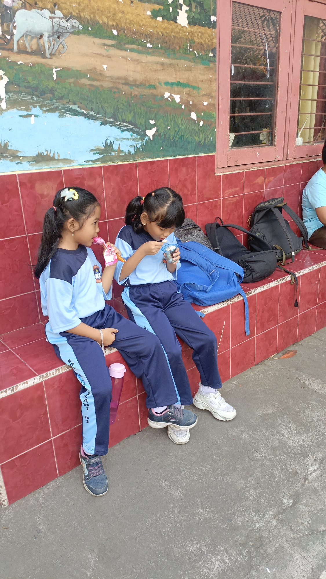 Two young girls, likely students given their matching light blue and dark blue uniforms, are sitting on a red tiled bench against a building. The building has a mural painted on the upper portion of the wall, depicting a pastoral scene with oxen plowing a field and a body of water. The lower portion of the wall is tiled in red. One girl on the left is holding a pink wrapper and appears to be eating from it. The girl on the right is holding a small cup and a spoon, also eating. They are positioned near a blue backpack and other bags on the bench. The ground in front of the bench is a concrete surface. The scene appears to be outdoors, possibly at a school. The mural, building style, and attire are consistent with locations in Salatiga, Indonesia.