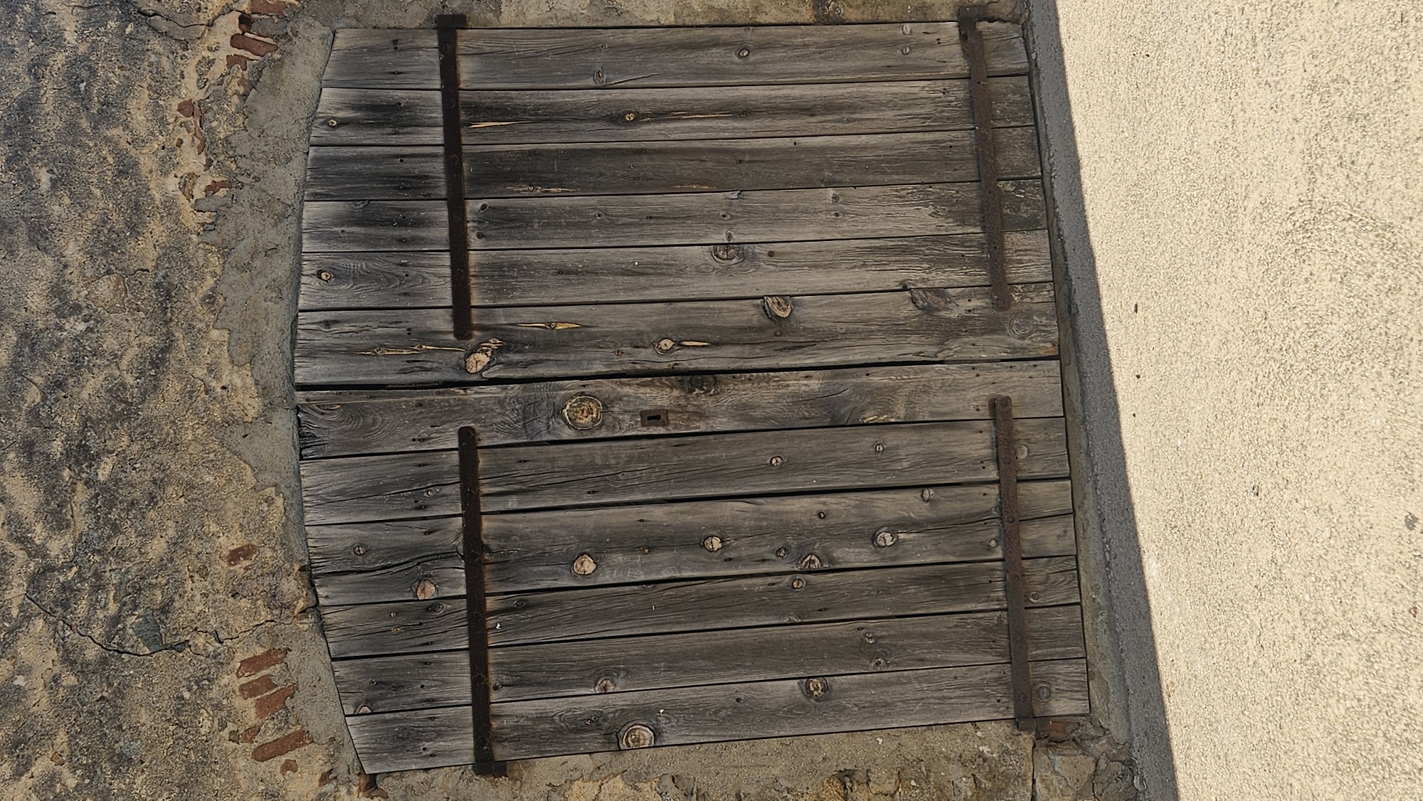 The image shows a pair of weathered wooden double doors set into a rough stone and stucco wall. The doors are made of vertical wooden planks with visible knots and grain, held together by horizontal metal straps that appear rusted. There is a keyhole visible on the left door, suggesting it is lockable. The wall above the doors is textured, with visible cracks and some exposed brickwork in the upper left corner. The ground in front of the doors is a light-colored, rough asphalt or concrete surface, with a distinct shadow cast from the doors. The overall impression is of an old, rustic entrance, possibly to a building or storage area in a Mediterranean or rural setting. The image is a still photograph with no people or animals present, focusing solely on the architectural elements. The context provided indicates this scene is located in L'Île-Rousse, France.