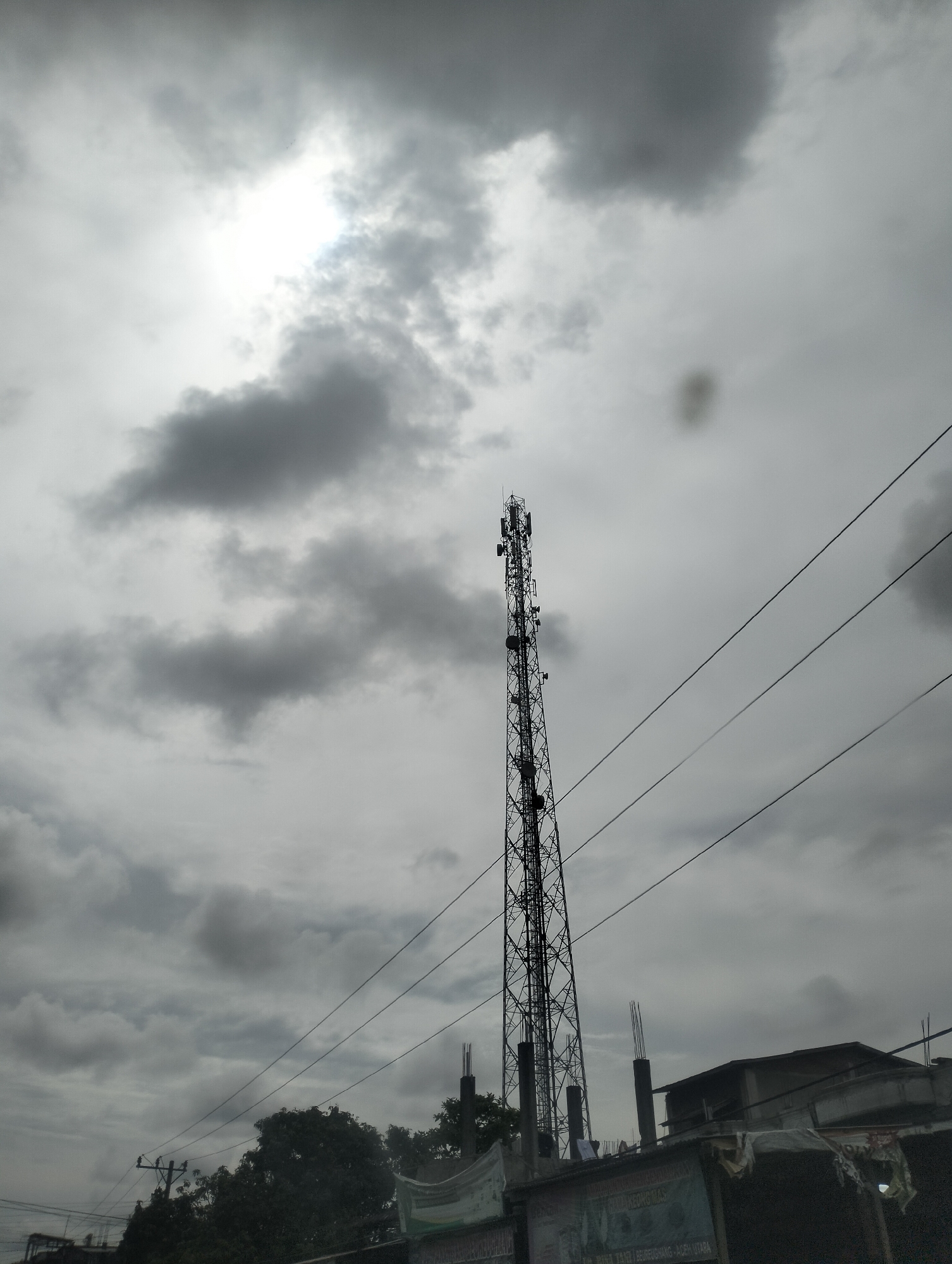 The image captures a slightly overcast sky over a city scene in Cotseutui, Indonesia. A tall, skeletal telecommunications tower dominates the center of the frame, adorned with multiple antennas and dishes. Several thick power lines stretch diagonally across the sky from right to left, appearing to pass near or over the tower. In the foreground and lower right of the image, a low-rise building structure is visible, partly obscured by the power lines. The building appears to have some construction elements, with visible rebar protruding from a concrete pillar. Some signage is present on the lower part of the building, though illegible due to distance and resolution. Trees and utility poles can be seen to the left and further in the background. The overall lighting suggests it is daytime, though the cloud cover diffuses the sunlight.
