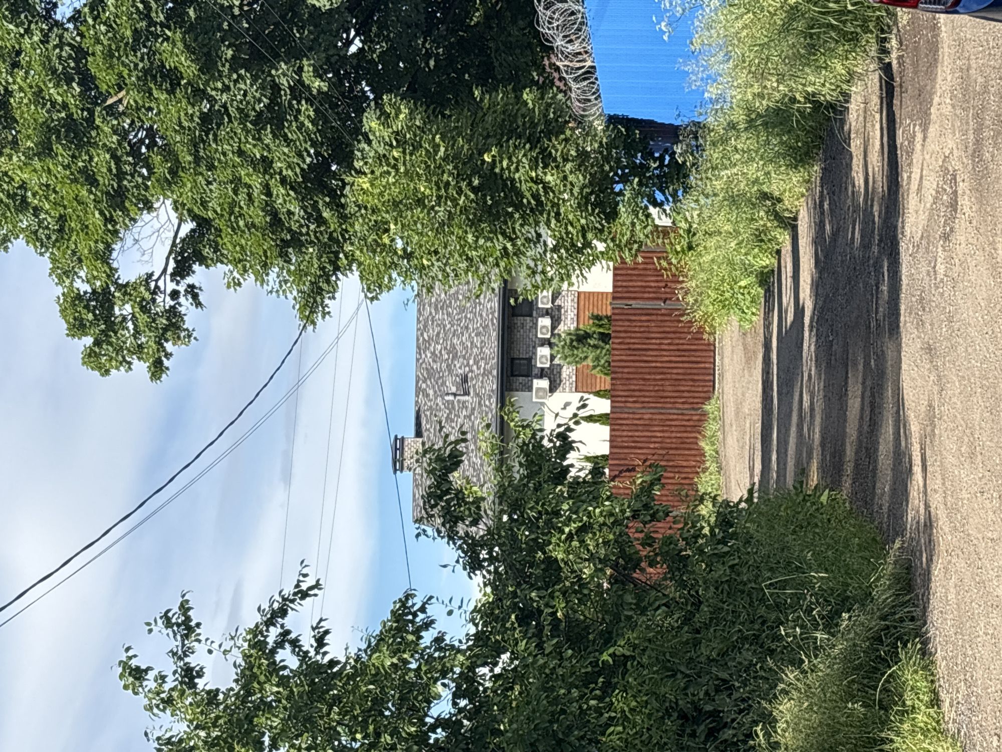 The image depicts a dirt path leading towards a house in Ostrov, Russia. The path is flanked by overgrown green vegetation on both sides. In the background, a house with a grey shingled roof and a brick chimney is visible. The house has several air conditioning units mounted on its facade. A reddish-brown wooden fence runs across the middle of the image, separating the path from the house. To the right of the house, a bright blue metal fence with barbed wire on top is present. The sky above is partly cloudy with a clear blue expanse. Power lines are strung across the sky. The overall scene suggests a rural or suburban residential area.