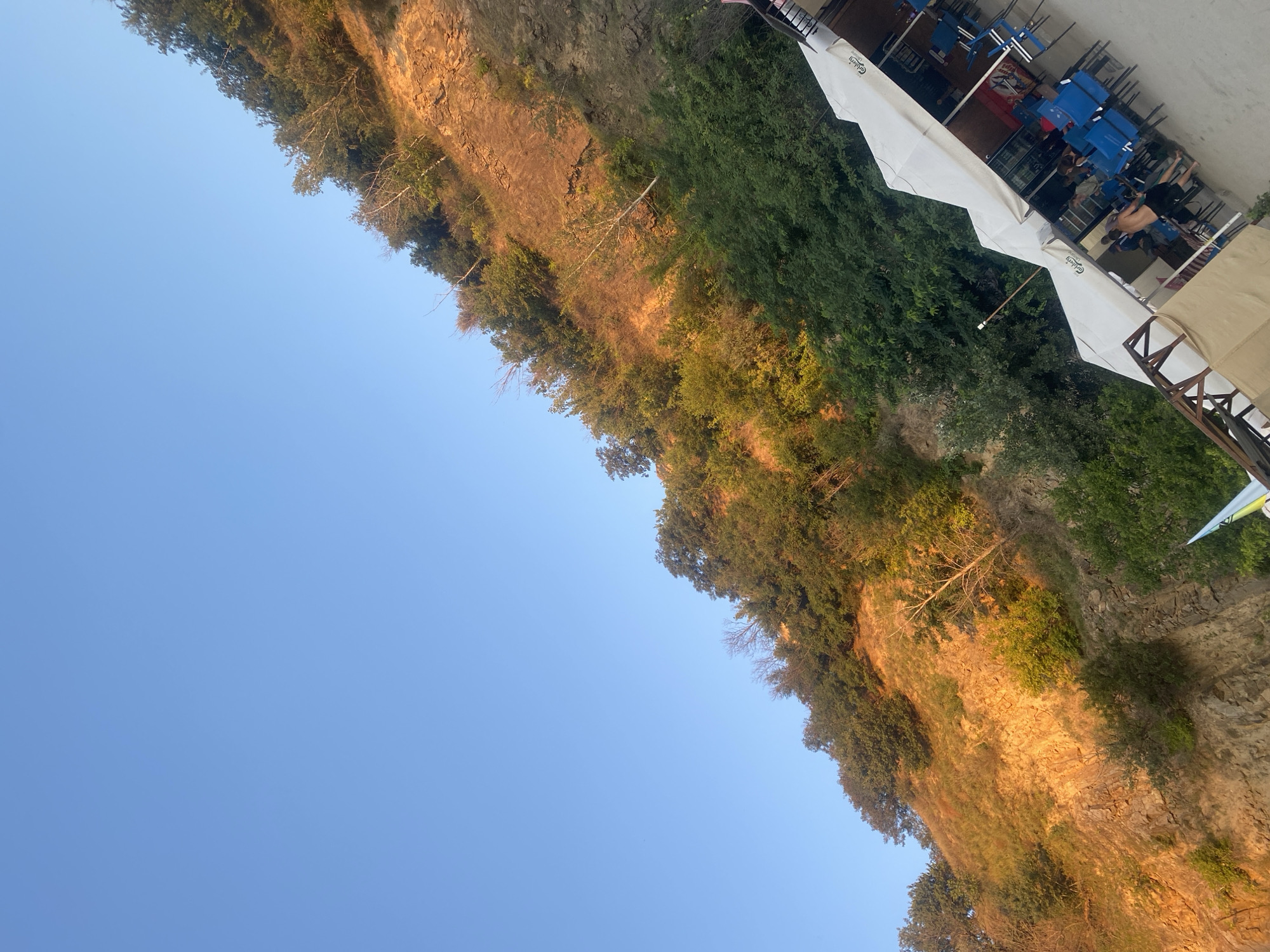 The image shows a cliff face covered in trees and vegetation, with a clear blue sky above. At the bottom right of the image, there are several white tents set up, resembling an outdoor cafe or seating area. Some blue chairs and tables are visible under the tents, and a few people are seated or standing around. The cliff is bathed in warm, golden light, suggesting it is either sunrise or sunset. The scene appears to be an outdoor recreational area, possibly a cafe located at the base of a cliff by a body of water, given the prevalence of such geological formations and establishments in areas like the Dnipro River embankment in Dnipro, Ukraine.
