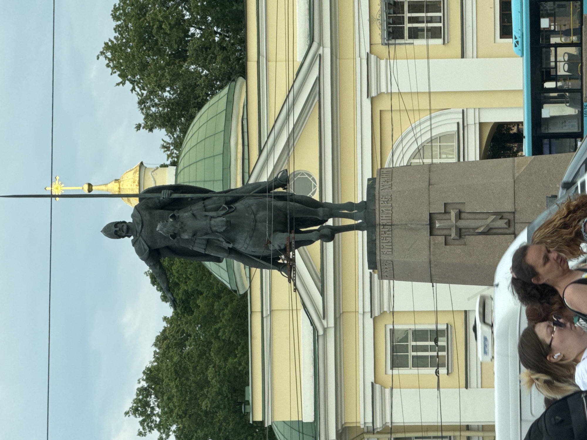 The media file shows a vertical outdoor scene dominated by a statue of a man on horseback, holding a spear. The statue is made of dark metal and is positioned in front of a pale yellow building with white architectural details and green domes. The building has several windows with white frames. There are also trees visible in the background and a bright blue bus on the right side of the frame. In the foreground, partially visible, are several people, some looking towards the statue or the surroundings. The sky appears overcast. The scene is likely in Saint Petersburg, Russia, as indicated by the architectural style and the presence of a statue often associated with historical figures. There is some visible text on the base of the statue, but it is not clearly legible. The overall impression is of a tourist site or a public square.