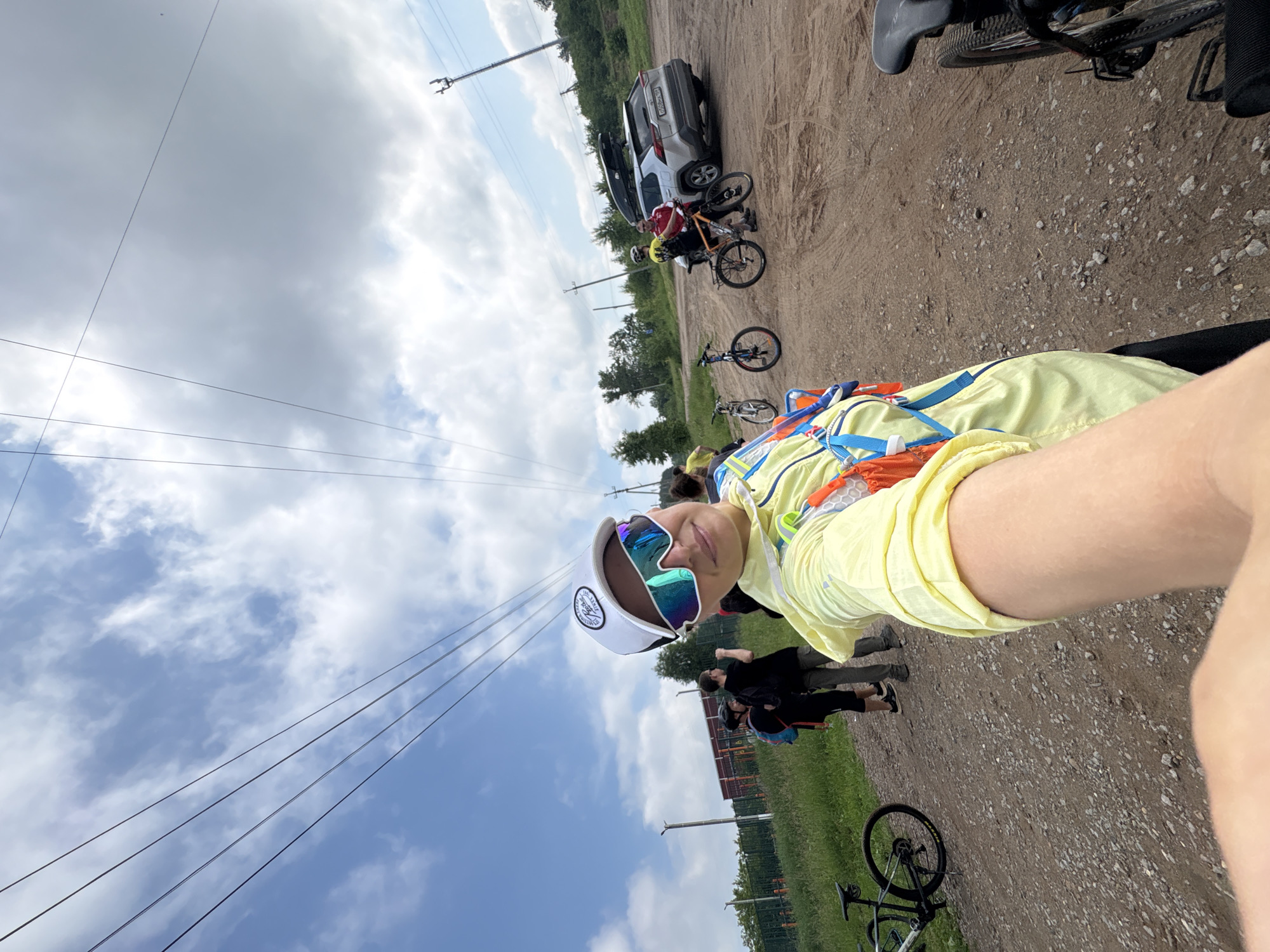In Kemul, Russia, on a gravel road under a cloudy sky, a group of people with bicycles are gathered. A woman in the foreground, wearing a yellow jacket and reflective sunglasses, takes a selfie. Behind her, several individuals are standing near bicycles. A silver SUV with a roof rack is parked nearby, and trees and a fence are visible in the background. Overhead power lines stretch across the sky. The scene suggests a cycling outing or event in a rural or semi-rural area.