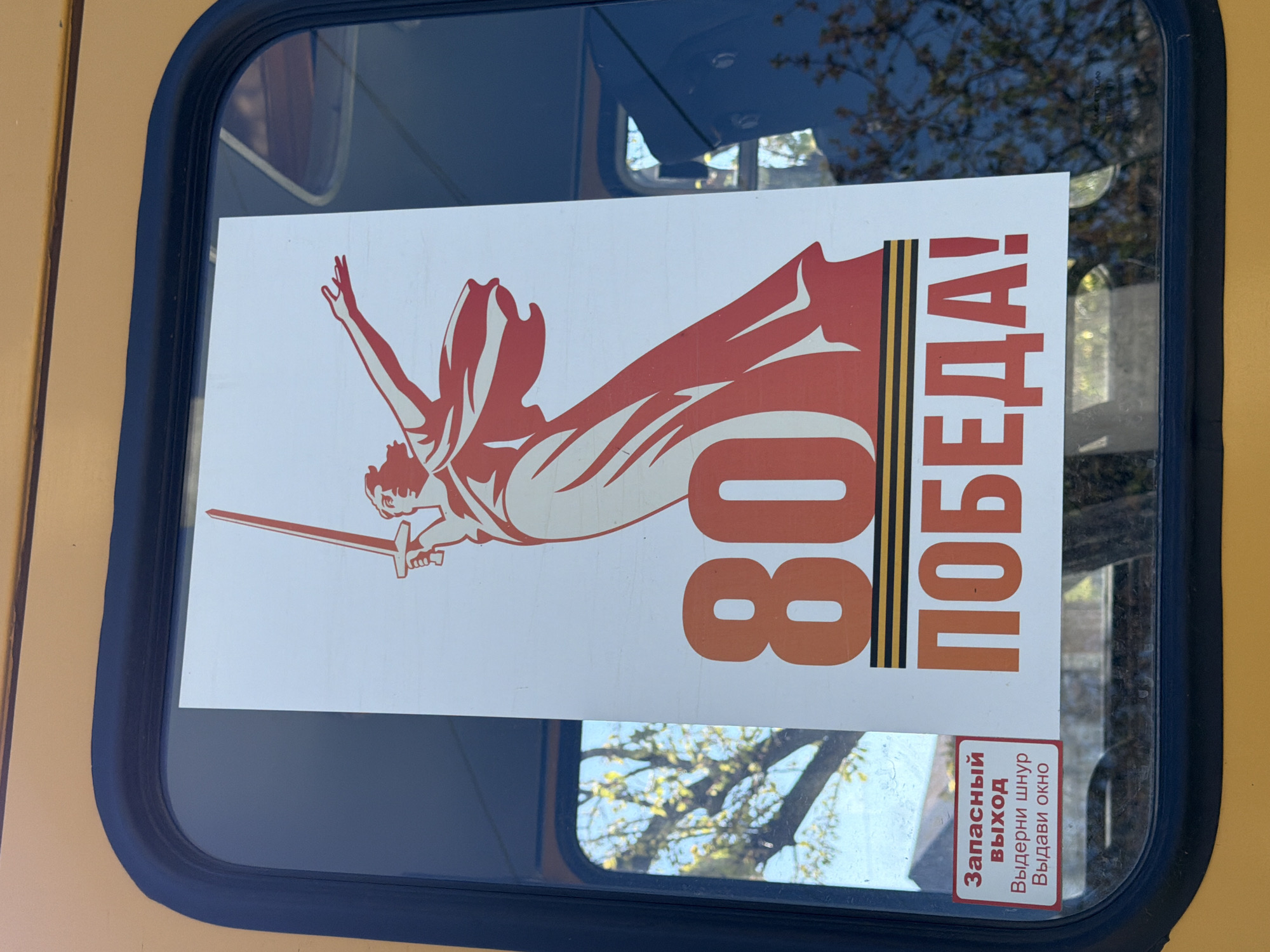 An image captures a white commemorative sticker affixed to the window of what appears to be a public transport vehicle in Yuzhny, Russia. The window has a dark blue frame, and the vehicle's exterior is a light yellow-orange color.
The main sticker features a stylized, reddish-orange graphic of a figure, reminiscent of the "Motherland Calls" statue, holding a sword and reaching upwards. To the right of the figure, large orange numbers "80" are visible. Adjacent to the numbers, vertically oriented, is the word "ПОБЕДА!" (Victory!) in red text. A prominent St. George's ribbon pattern, with three black stripes and two orange stripes, vertically separates the "80" from "ПОБЕДА!". This iconography strongly suggests a commemoration of Victory Day, likely an 80th-anniversary celebration of the victory in World War II.
Reflections on the window show parts of a blue sky and green-leaved trees, indicating that the photo was taken outdoors during daylight hours.
In the bottom right corner of the window, a smaller red-and-white rectangular sticker is visible. It contains emergency instructions in Russian: "Запасный выход" (Emergency Exit), "Выдерни шнур" (Pull cord), and "Выдави окно" (Push out window), further confirming the setting as a vehicle.