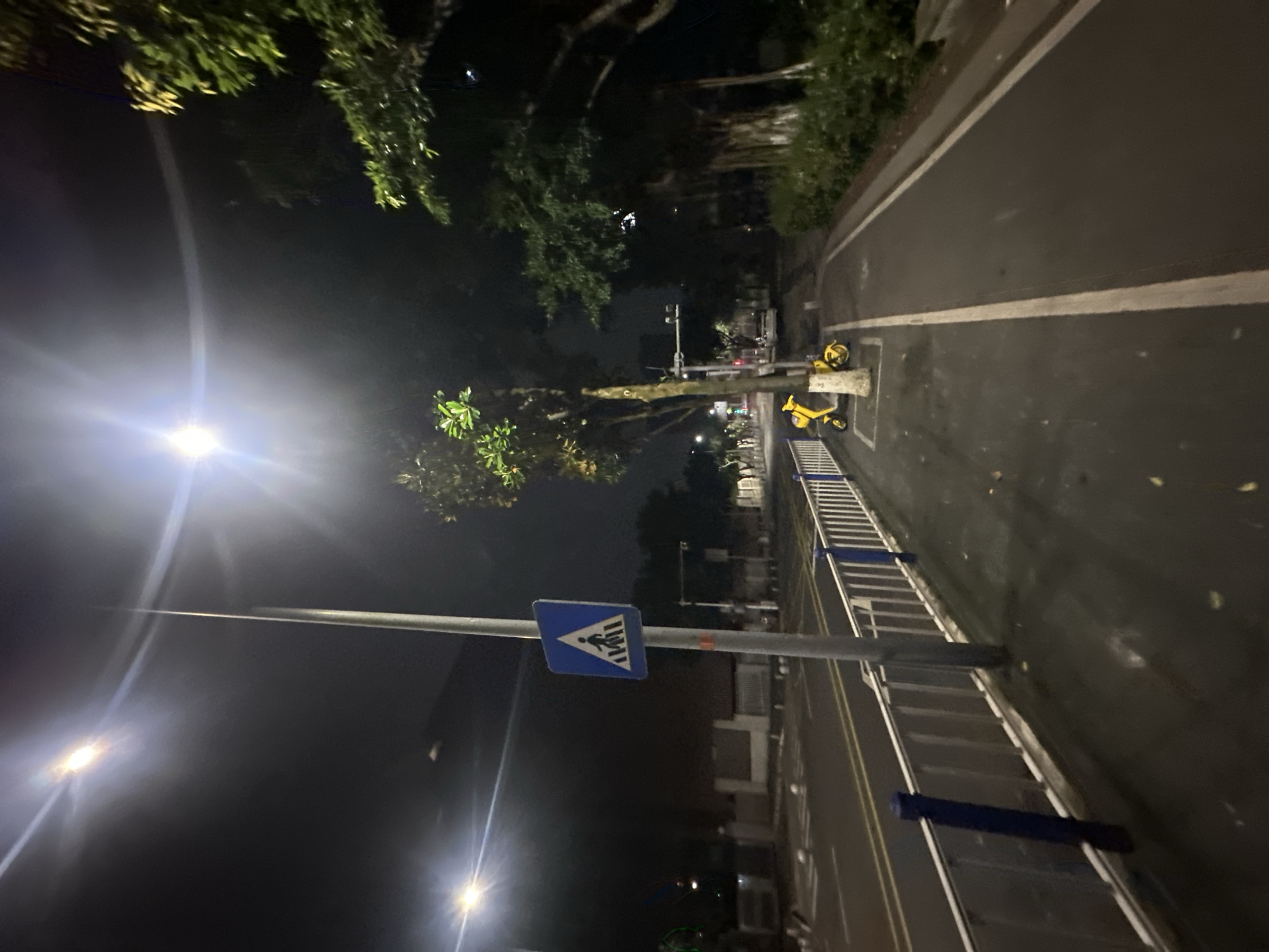 The image captures a street scene at night in Chongqing High-tech Zone, China. A pedestrian crossing sign is visible on a light pole on the left side of the frame. A metal fence with blue posts separates the sidewalk from the road. Two yellow shared electric scooters are parked on the sidewalk. The road is empty except for a white car in the distance. Trees line the right side of the sidewalk, and streetlights illuminate the scene. The overall environment suggests a quiet urban street.
