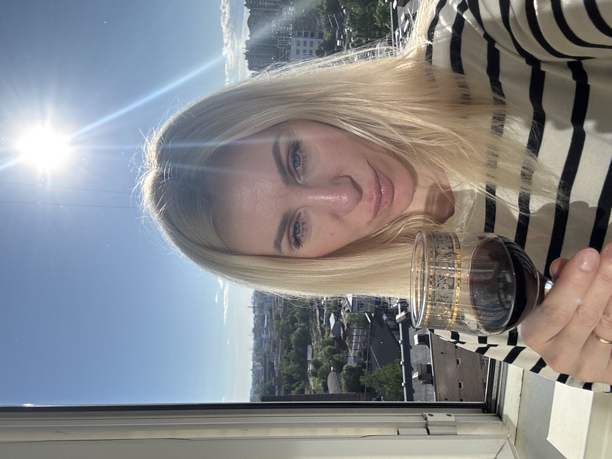 A blonde woman is pictured in a selfie, holding a glass of red wine and looking at the camera. She is wearing a striped shirt. The background shows a cityscape under a bright sun and blue sky, with some clouds. The woman appears to be indoors, near a window, with a view of buildings and trees.