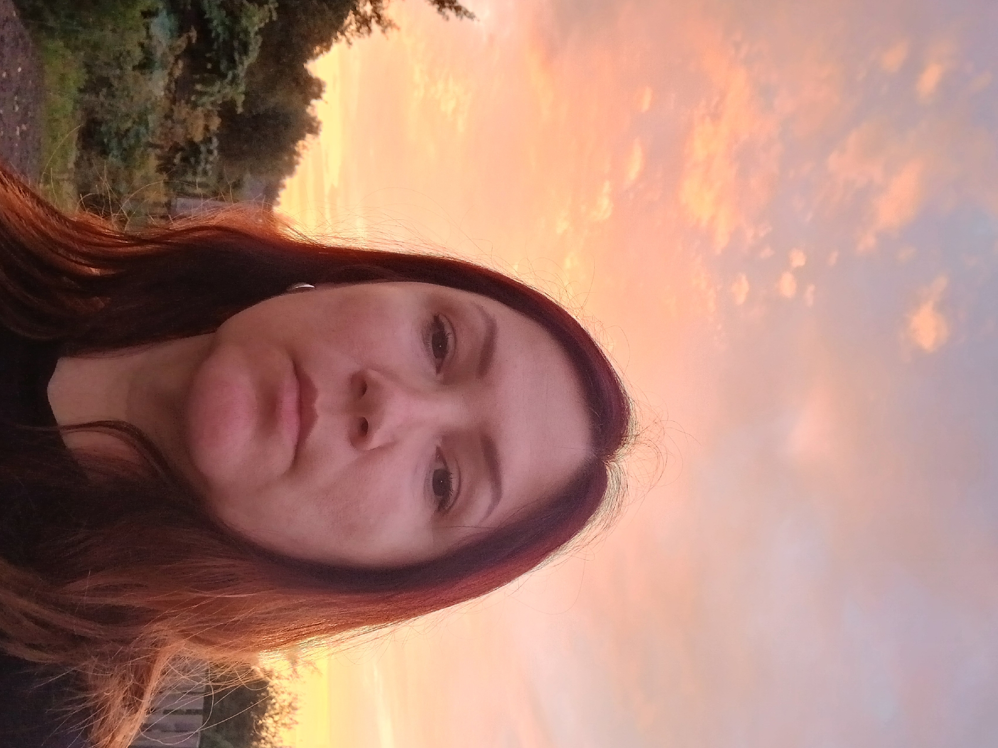 An outdoor, close-up, eye-level, full-face, self-portrait captures a woman from the chest up against a vibrant, orange and pink sunset sky filled with wispy clouds. The woman has medium-toned skin, dark brown hair with reddish highlights, and is looking directly at the camera. She is wearing a dark-colored shirt and a small silver hoop earring in her visible left ear. Behind her, a blurred background shows the silhouettes of trees and vegetation, suggesting a rural or natural setting. The lighting suggests it is dusk or dawn.