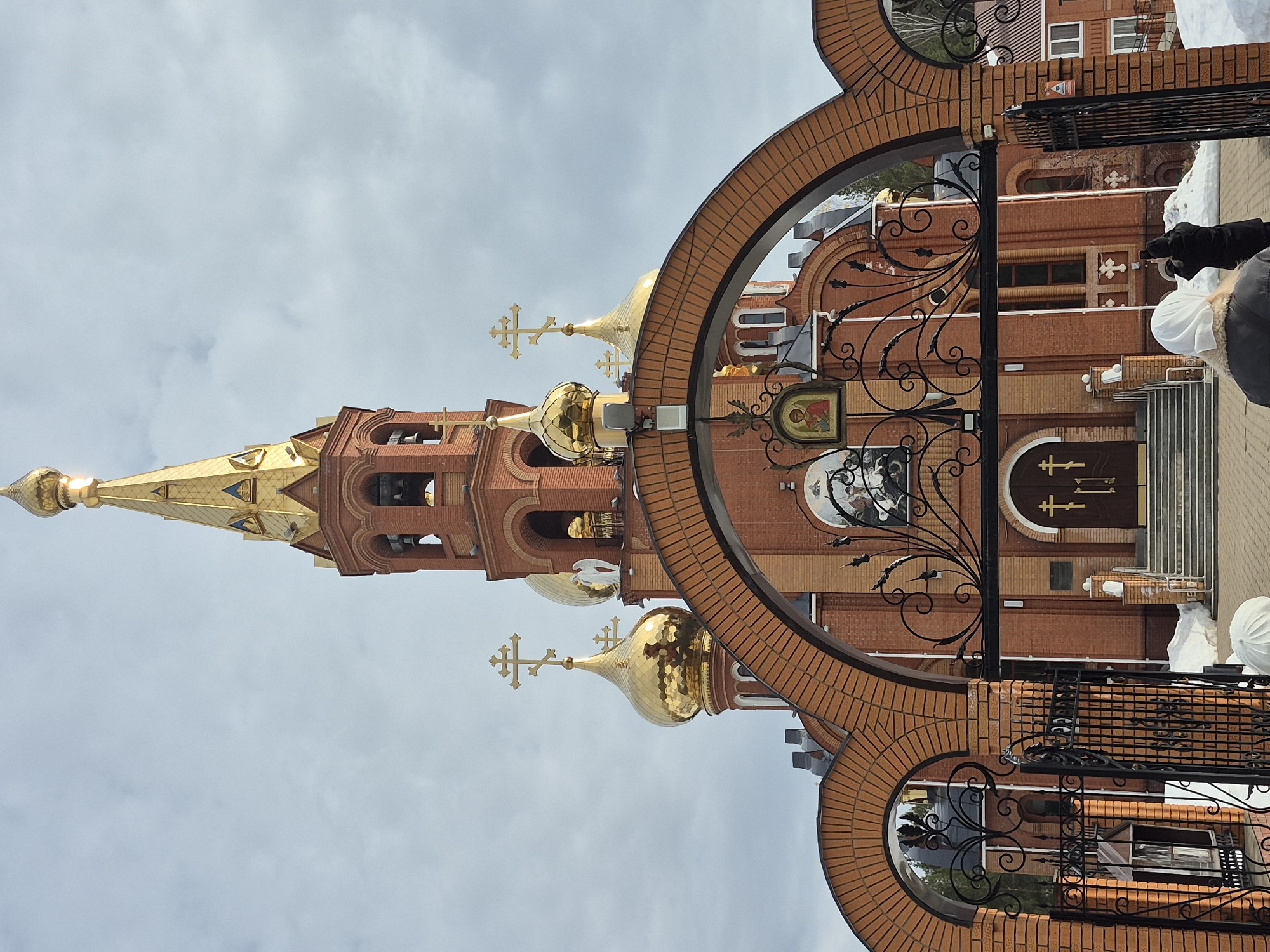 The image depicts the Church of St. George the Victorious, located at ulitsa Karla Marksa, 47, in Chaikovsky, Perm Krai, Russia. The church is a large, multi-domed brick structure with a prominent central bell tower topped by a golden spire and an Orthodox cross. Several other golden onion domes, also crowned with golden crosses, are visible across the church's roofline. The facade is constructed of red brick with white trim and features arched windows. Religious iconography and murals are present on the exterior walls, including a large mural depicting figures on horseback above the main entrance.
In the foreground, a substantial brick archway serves as an entrance, with open black wrought-iron gates featuring decorative scrollwork. A small icon of a saint is affixed to the gate structure. Two individuals are positioned from behind, below this archway, facing the church. One person in the immediate foreground wears a white headscarf and a dark coat. Behind them, another individual in a dark coat holds a smartphone, apparently photographing the church.
The ground leading to the church entrance is paved, while patches of snow are visible on either side of the path, suggesting a cold season. The sky overhead is overcast with light grey clouds. The main entrance to the church consists of double brown doors adorned with crosses, accessible via a set of stairs. A white statue resembling an angel is situated near one of the golden domes on the church's upper structure.