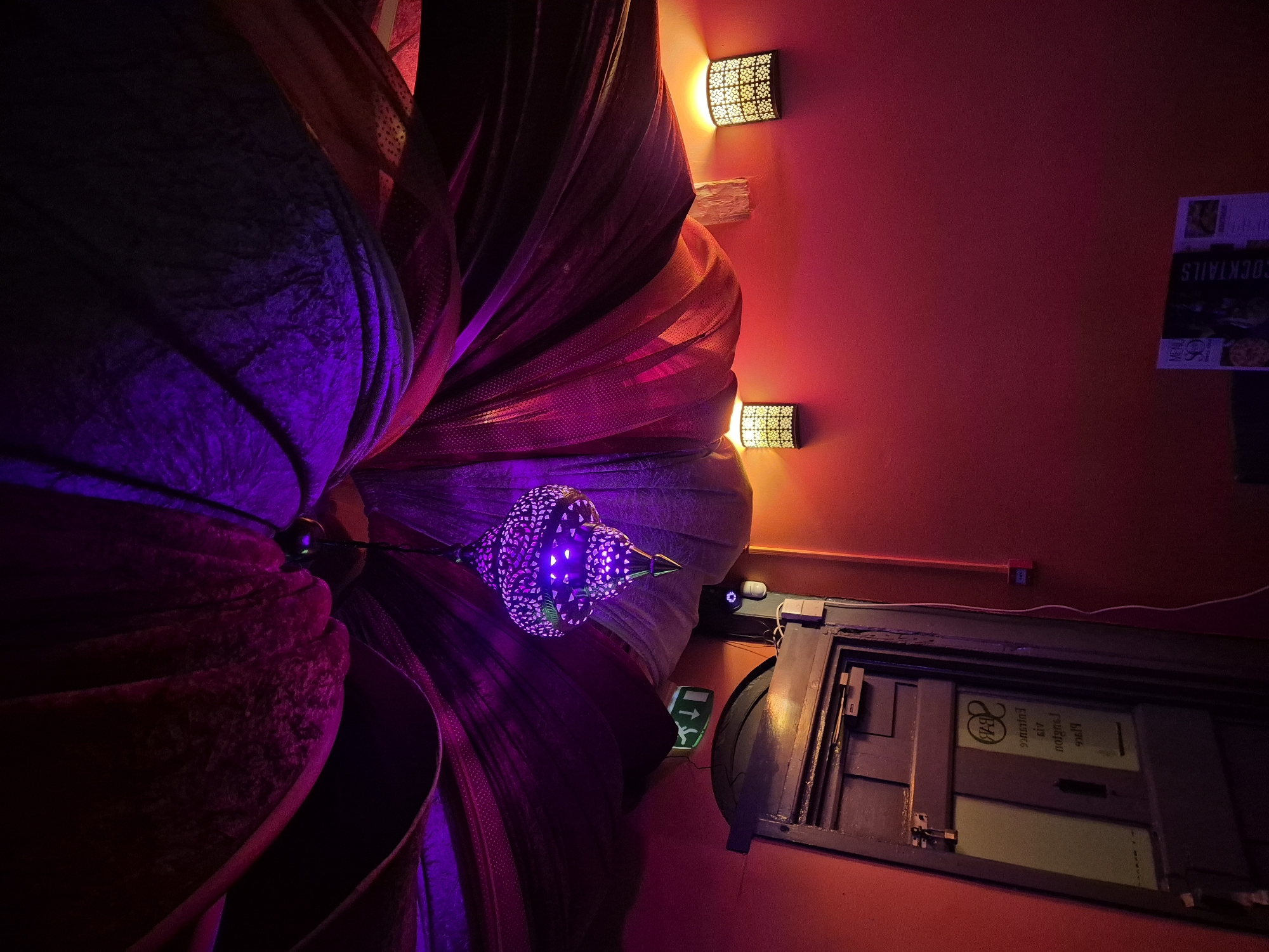 The image, taken from a low angle in what appears to be an interior space in West Suffolk, United Kingdom, shows a ceiling adorned with decorative elements. A large, flower-like fabric arrangement is the dominant feature, illuminated with purple and red lighting. Hanging beneath it is a smaller, decorative lamp with a patterned design, also lit in purple.
The ceiling is painted a dark red. Two decorative wall sconces with floral-patterned cutouts provide ambient lighting. To the right, there's an emergency exit sign with a green arrow pointing to the left. Below it is a framed display with text and images that appear to be promotional material, partially obscured by the darkness. There is also a door in the lower right corner.