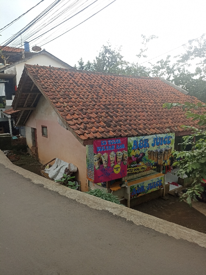 The image shows a small, one-story building with a red tile roof, located in Jatinangor, Indonesia (latitude: -6.936, longitude: 107.779). The building has a white facade with a small window and a sign advertising "Es Teh Susu dan Air" and "Air Juice." The sign is on a wooden frame, with a pink background, and displays pictures of various beverages. The building is situated on a slope, and there is a concrete curb separating the building from a paved road in the foreground. There are plants and shrubs growing around the building. The sky is overcast, with power lines running across the top of the image. The image is taken from a slightly elevated angle, looking down at the building.