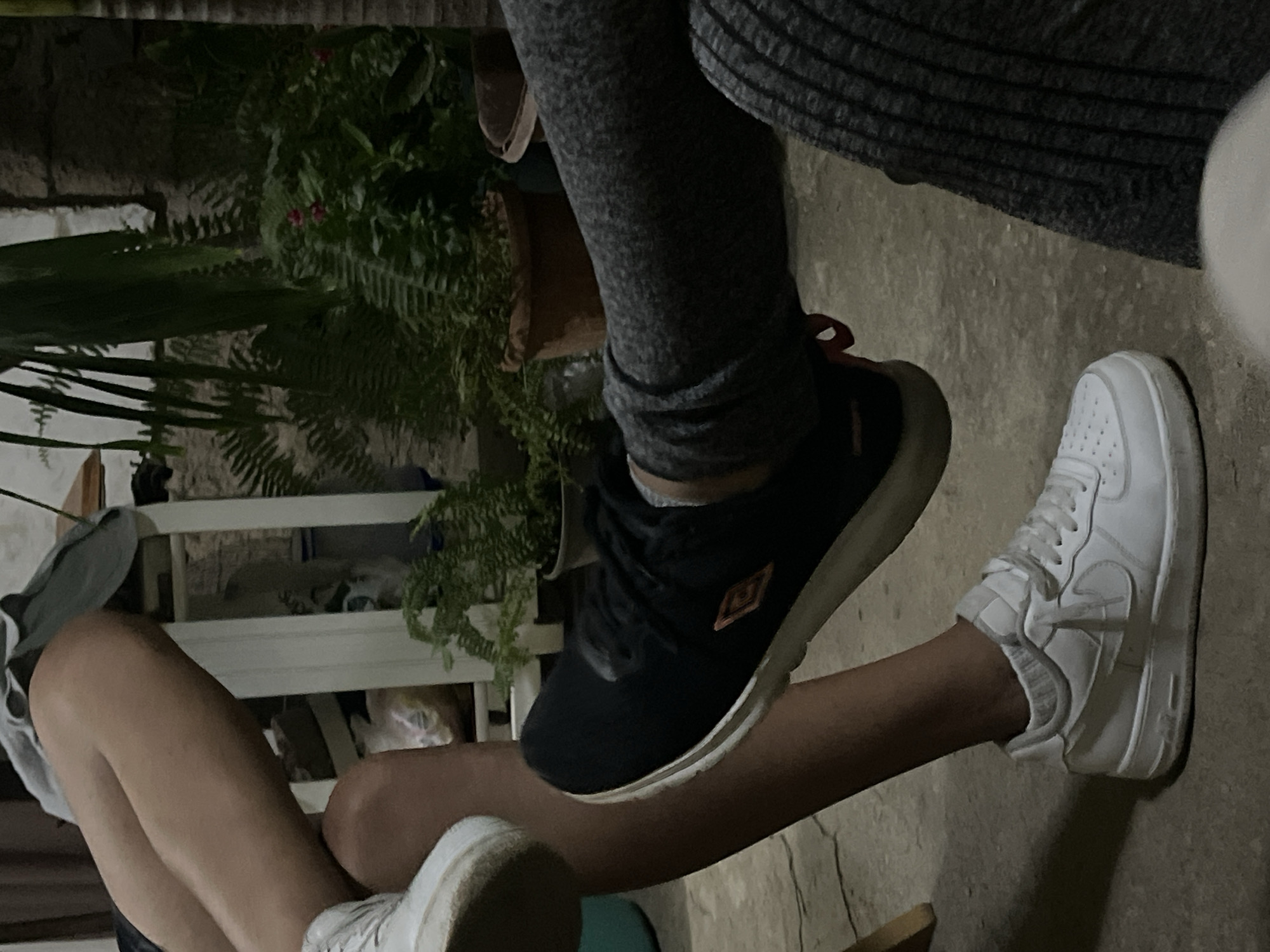The image shows a close-up of two people's legs and feet, with one person wearing white sneakers and the other wearing black sneakers. They appear to be sitting outdoors on a concrete surface, with some potted plants and a stone wall in the background. The lighting suggests it may be evening or nighttime. The scene is likely a residential area in Santa Ana, Venezuela, given the visual cues of common outdoor living spaces in such a location.