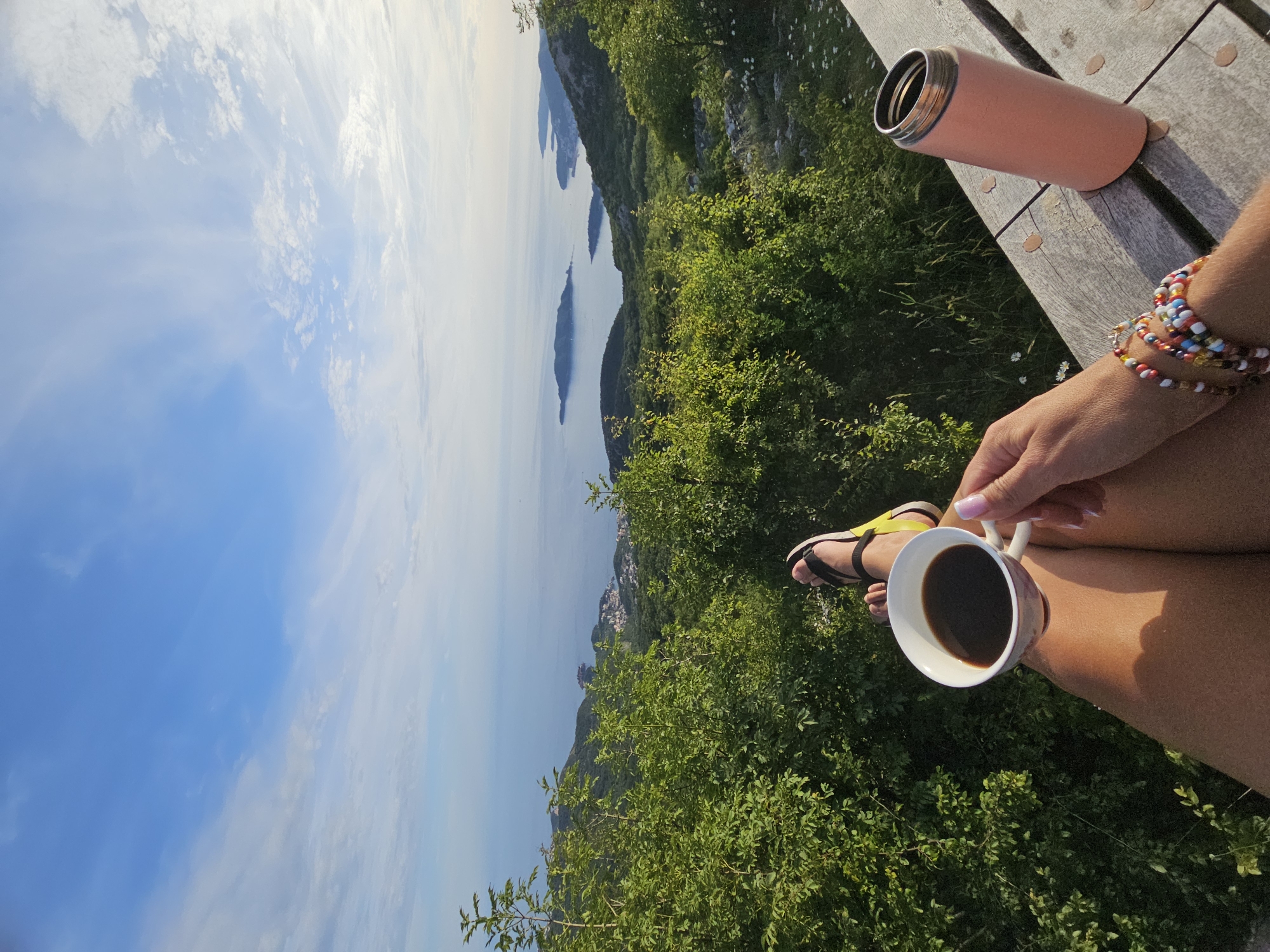 The image, taken from a first-person perspective, captures a person relaxing at a high vantage point overlooking a scenic coastal area. The setting is a high vantage point overlooking a coastal landscape, likely a private terrace or viewpoint, located in the city of Unknown, Unknown.
In the foreground, the person's tanned legs and feet are visible, resting on a light-colored wooden surface, possibly a deck or table. The person wears black and yellow open-toed sandals. Their right hand, adorned with light pink nail polish, holds a white ceramic mug filled with dark liquid, presumably coffee. The mug has a subtle floral or patterned detail on its lower portion. To the left of the person's arm, which is decorated with multiple colorful beaded bracelets, a pink insulated bottle with a silver rim stands upright on the wooden surface. The wooden surface exhibits grain and a few small, scattered, light-colored circular marks, possibly from fallen leaves or minor imperfections.
Immediately beyond the wooden surface and stretching into the mid-ground, dense green foliage, consisting of various bushes and small trees, dominates the hillside. Further in the mid-ground, a panoramic view unfolds: a calm body of water, likely the sea or a large bay, is visible. Several green, treed islands or peninsulas protrude from the water. On the left side of the coastline, nestled along the shore, a compact town or village with numerous buildings can be seen.
The background features a vast, clear blue sky with sparse, wispy white clouds scattered across it. The sky transitions from a slightly lighter hue near the horizon to a deeper blue overhead. The lighting suggests either early morning or late afternoon, casting soft, warm tones and creating a serene atmosphere.