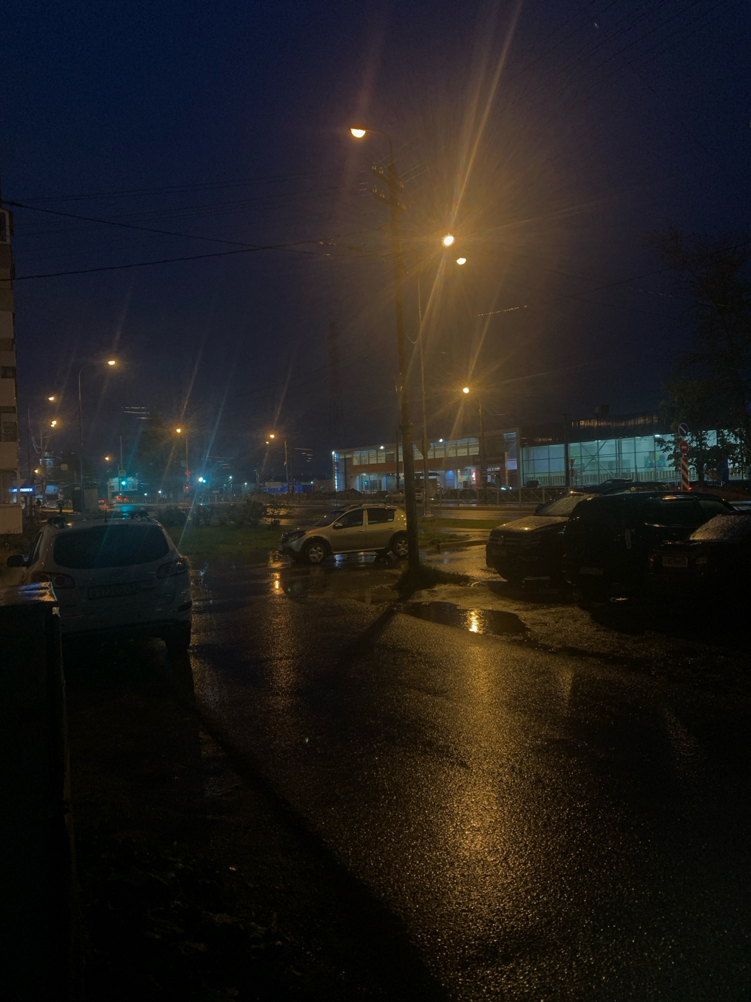 A low-angle, medium shot captures a street scene in Arkhangelsk, Russia at dusk. The sky is a deep, dark blue, illuminated by the warm, yellow glow of streetlights and light from buildings. The wet asphalt of the street reflects these lights, creating streaks of yellow and orange. Several cars are parked along the street; a white SUV is parked on the left, facing away from the camera, and a dark SUV is parked in the center, facing towards the camera. A lighter-colored car is parked to the right of the dark SUV. In the background, a large building with illuminated windows is visible, suggesting a commercial area. The overall atmosphere is one of a quiet, damp evening in a Russian city.