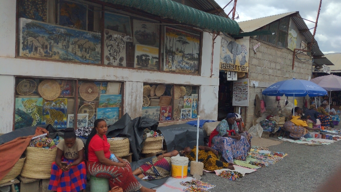This image depicts an outdoor market scene identified by a prominent sign as "SERENGETI STREET ENTRY" in Moshi, Tanzania. The scene is dominated by a light-colored building on the left, featuring display windows filled with various paintings—including large depictions of leopards, village scenes, and landscapes—alongside numerous woven baskets and decorative plates. Adjacent to this building, a structure of grey concrete blocks is visible.
In the foreground, three adult women are present. On the left, a woman in a red and blue plaid skirt and a light top sits on a low surface, engaged with a mobile phone. To her right, another woman in a red top and patterned skirt sits on a small stool, facing right. Behind them, several large, woven cylindrical baskets are stacked. Further right, a woman in a patterned dress lies flat on a dark fabric spread directly on the unpaved ground, with a yellow plastic bucket positioned next to her head.
Further into the scene on the right, several other women are seated on the ground beneath a blue market umbrella and a lighter umbrella. They are surrounded by various small colorful items, primarily appearing to be beaded jewelry and fabric crafts, spread out on cloths. The ground is an unpaved surface of compacted dirt or gravel. The sky is overcast.