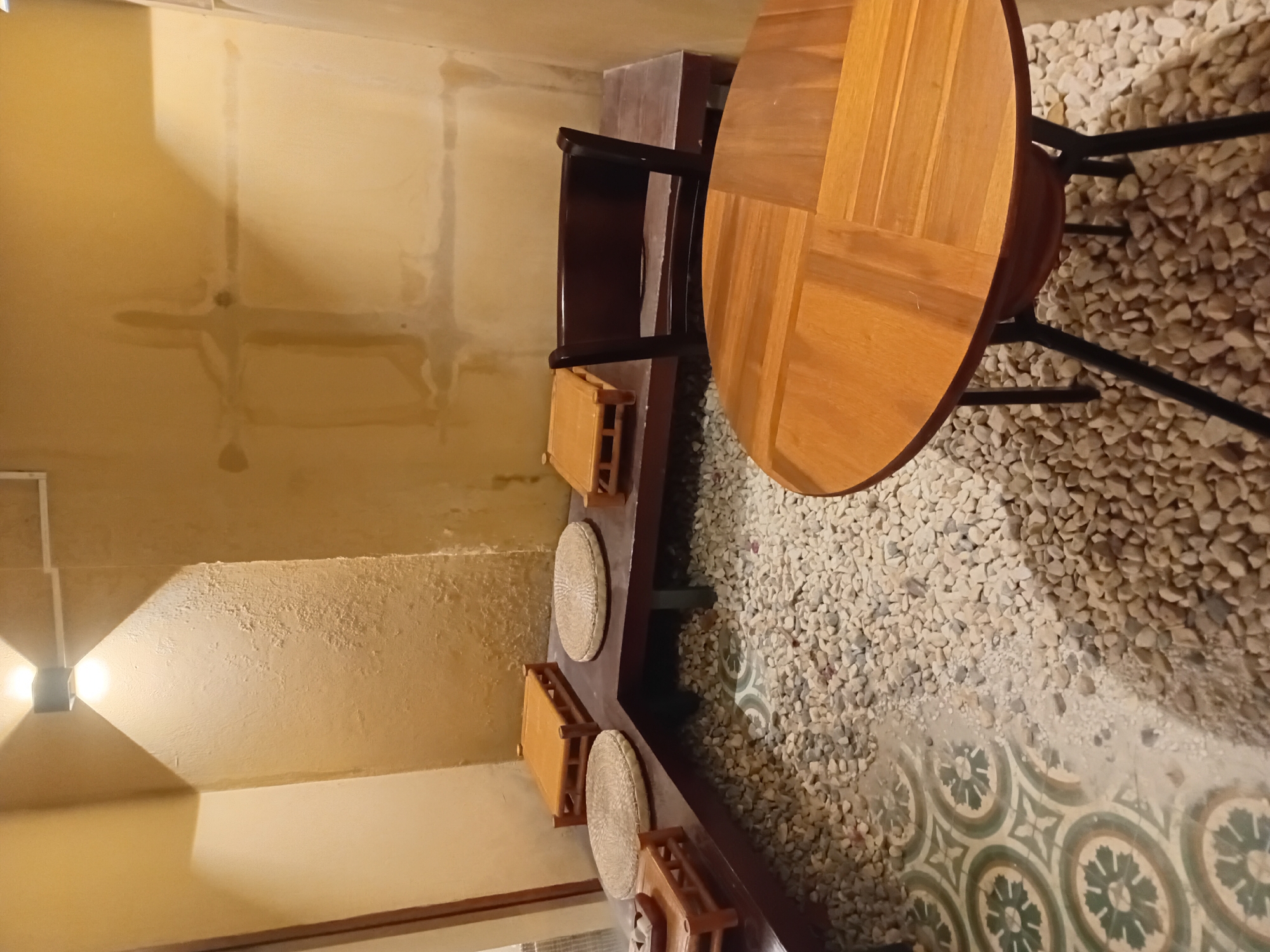 The image depicts a cozy seating area with a Japanese aesthetic, likely a cafe or restaurant. The floor is covered with small, light-colored pebbles, with a section of decorative tiled flooring visible on the left. A low, dark wooden bench runs along the back wall, topped with several square wooden seats and round woven cushions. In front of the bench, a dark wooden chair is placed at a circular, light-wood table. The walls are a textured, muted yellow, and a modern wall lamp illuminates the upper corner. The overall impression is one of tranquility and minimalist design, suggestive of a peaceful retreat within the city of Đà Nẵng, Vietnam.
