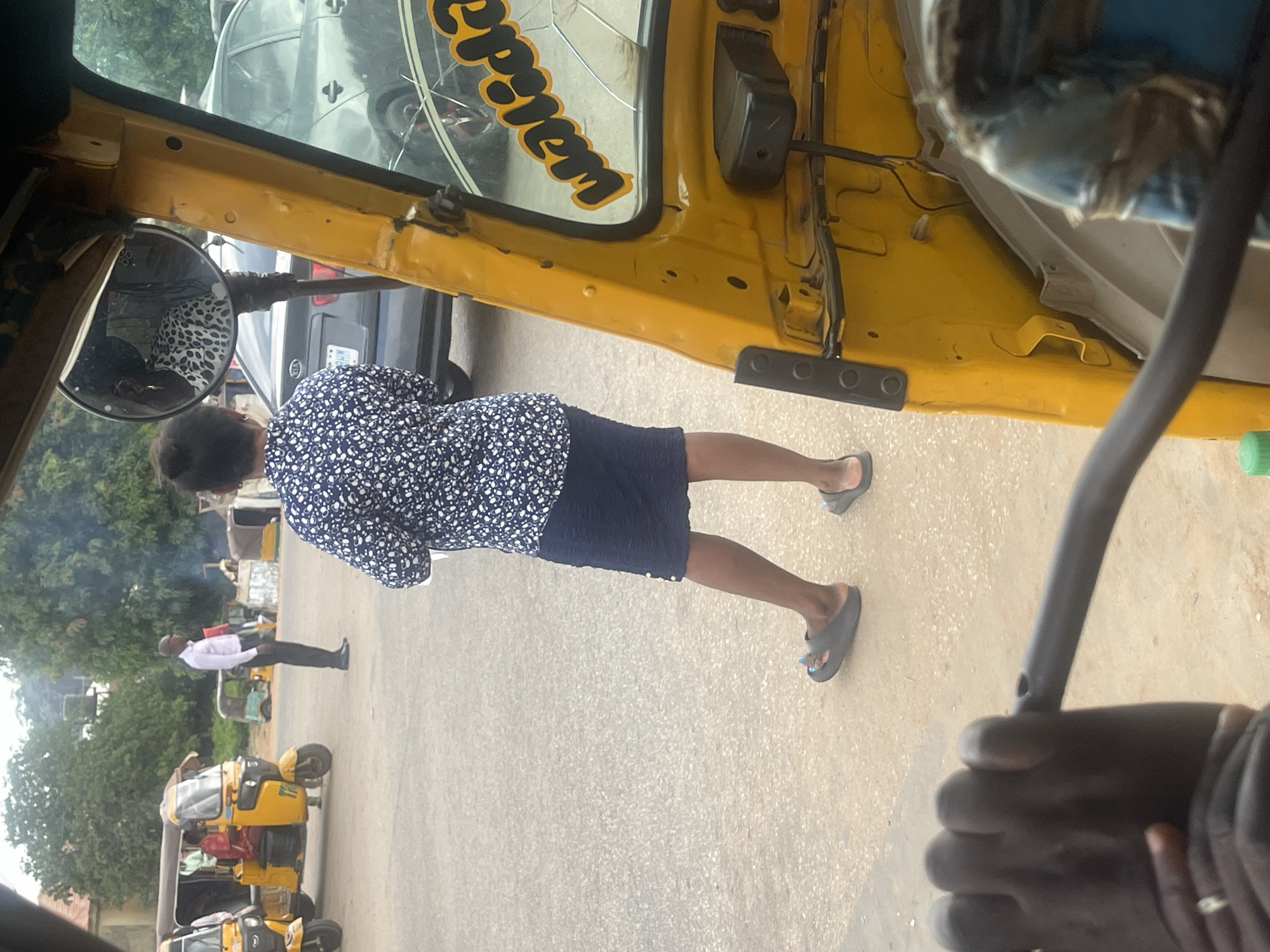 A medium shot shows a woman in a navy blue and white floral print top and a dark blue skirt, standing with her back to the camera in a dusty outdoor area. She is wearing grey slip-on sandals. To her left, a man in a white shirt and black pants stands facing away from the camera, and a yellow auto-rickshaw is parked. In the background, other auto-rickshaws and a silver sedan are visible. The foreground shows the interior of a yellow auto-rickshaw, with a dark handle bar and the hand of the person operating it visible. The scene appears to be a street or transport hub in Galadimawa Village, Nigeria, characterized by unpaved ground and several vehicles.