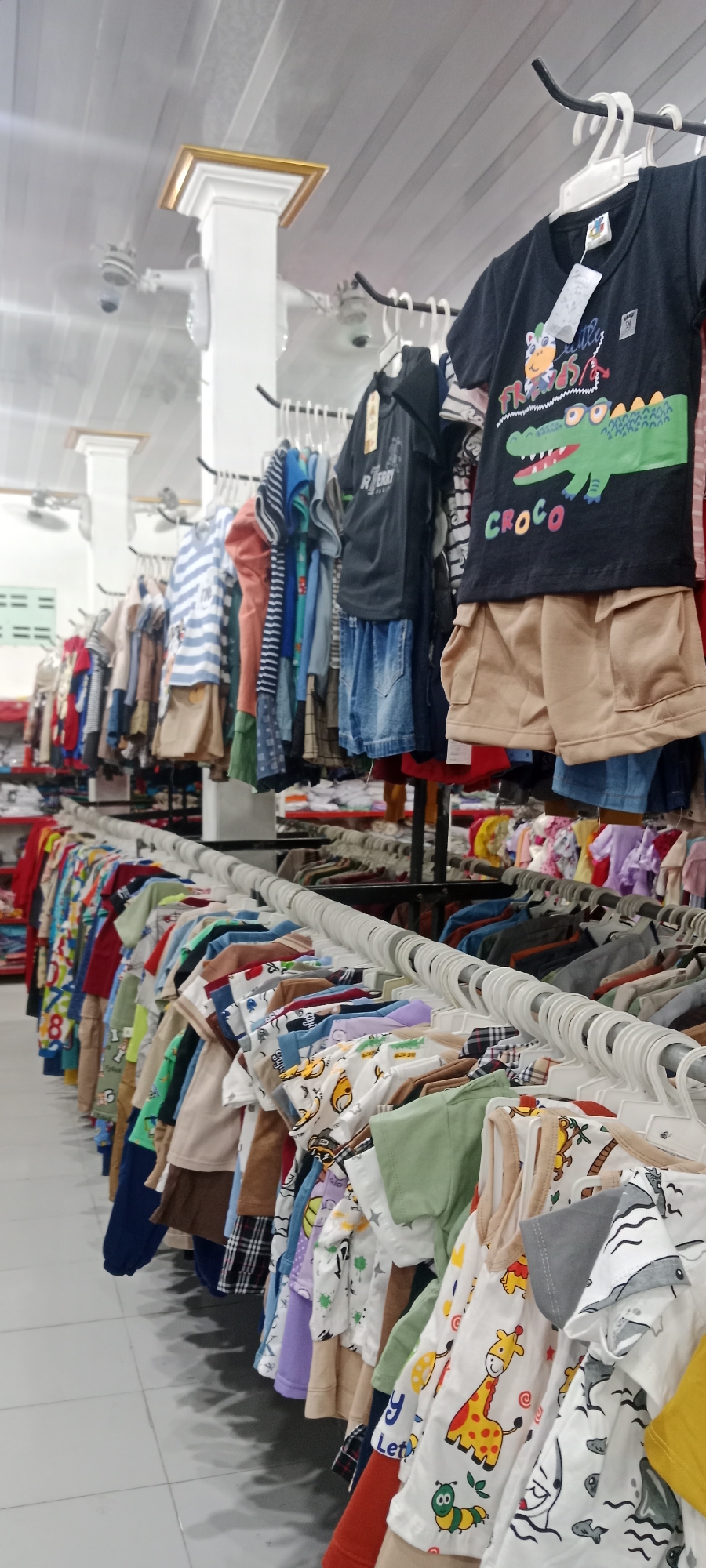In the city of Rengel, Indonesia, an indoor retail store features a well-lit display of children's clothing organized on hanging racks. The clothing is predominantly t-shirts and shorts, with a variety of colors and patterns including stripes, checks, and cartoon graphics like a crocodile and a giraffe. The racks are arranged in rows, extending into the background, suggesting a substantial inventory. The store's interior is clean and organized, with tiled flooring and white walls. Overhead lighting illuminates the merchandise. Two white ceiling fans are visible in the upper portion of the image, along with decorative white pillars topped with gold trim. There are no people visible in the frame.