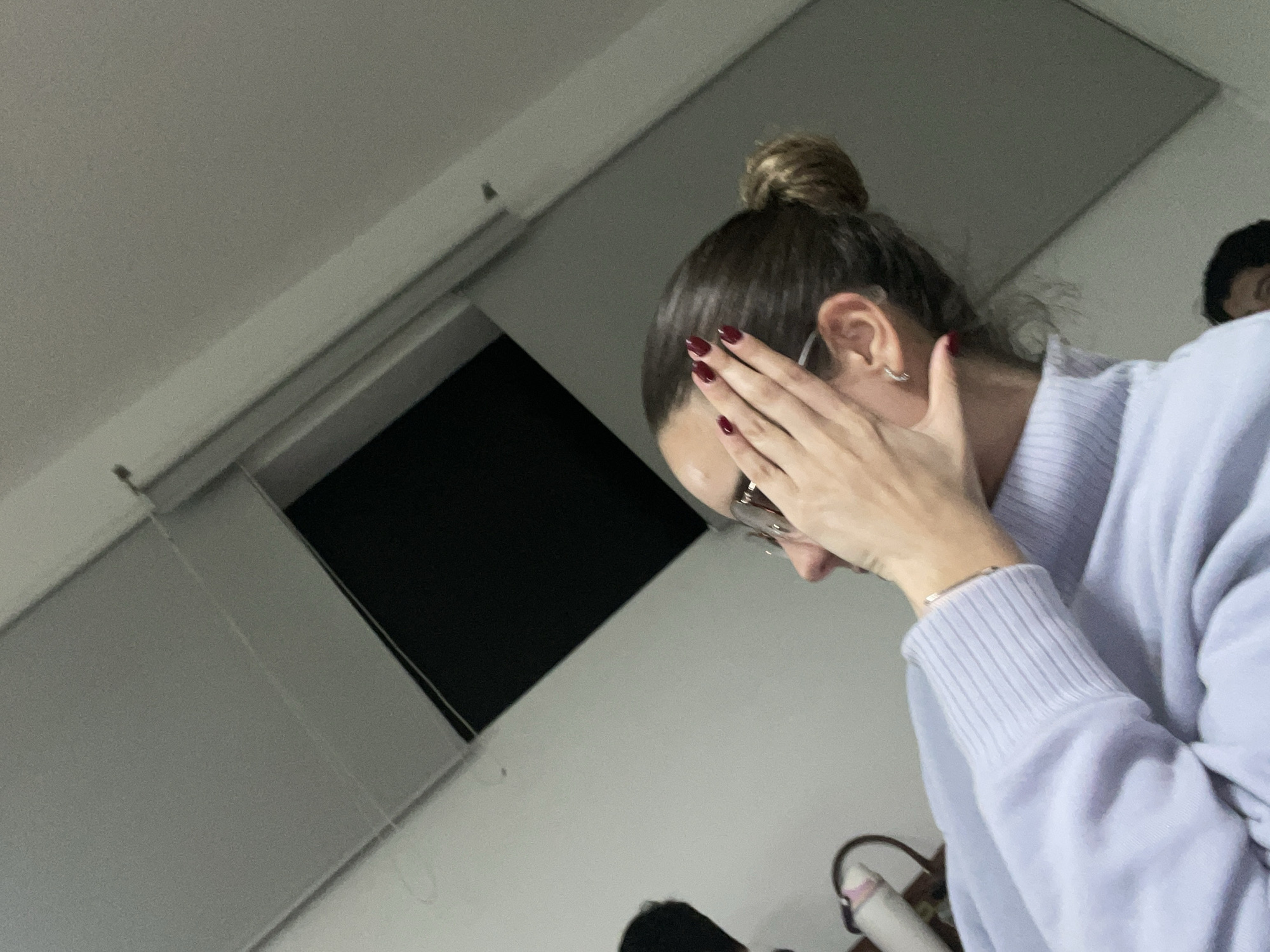 The image depicts an indoor scene in San Andrés Cholula, Mexico, featuring a female subject partially obscuring her face and another individual in the background. The primary subject is a female, seen in profile, facing towards the right. Her dark brown hair is styled in a messy bun. She wears clear-framed glasses and a light lavender turtleneck sweater with ribbed cuffs. Her right hand is raised, with fingers slightly splayed, covering the right side of her face from the eye downwards. Her fingernails are painted dark red. A thin silver bracelet is visible on her right wrist, and a silver hoop earring is in her right ear. In the lower right corner, the top of a second individual's head, with dark hair, is partially visible. The background consists of light-colored walls and ceiling. A dark, rectangular void, possibly an open window or doorway to an unlit area, is present in the upper left and center-right portions of the frame, bordered by light-colored framing. To the left of the primary subject's head, a partially visible, indistinct light-colored object, possibly a bag or bottle, is present. The lighting is soft and ambient.